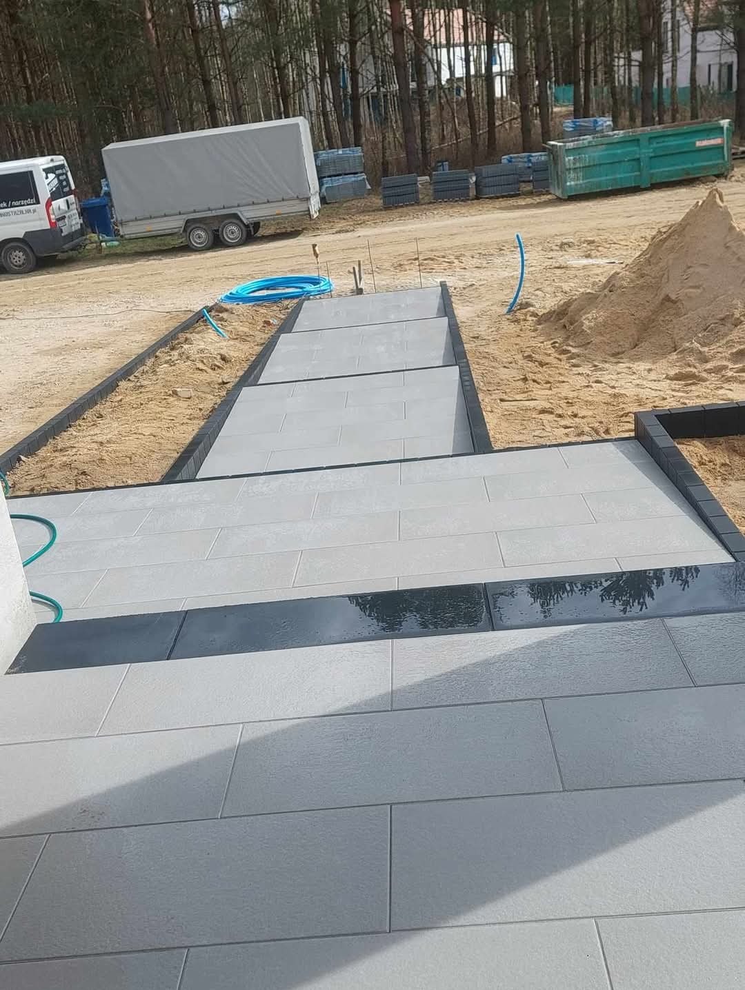 Ułożone schody z jasnoszarej kostki brukowej z czarnymi obrzeżami na posesji. W tle widoczny piasek, kontener i samochód dostawczy.
