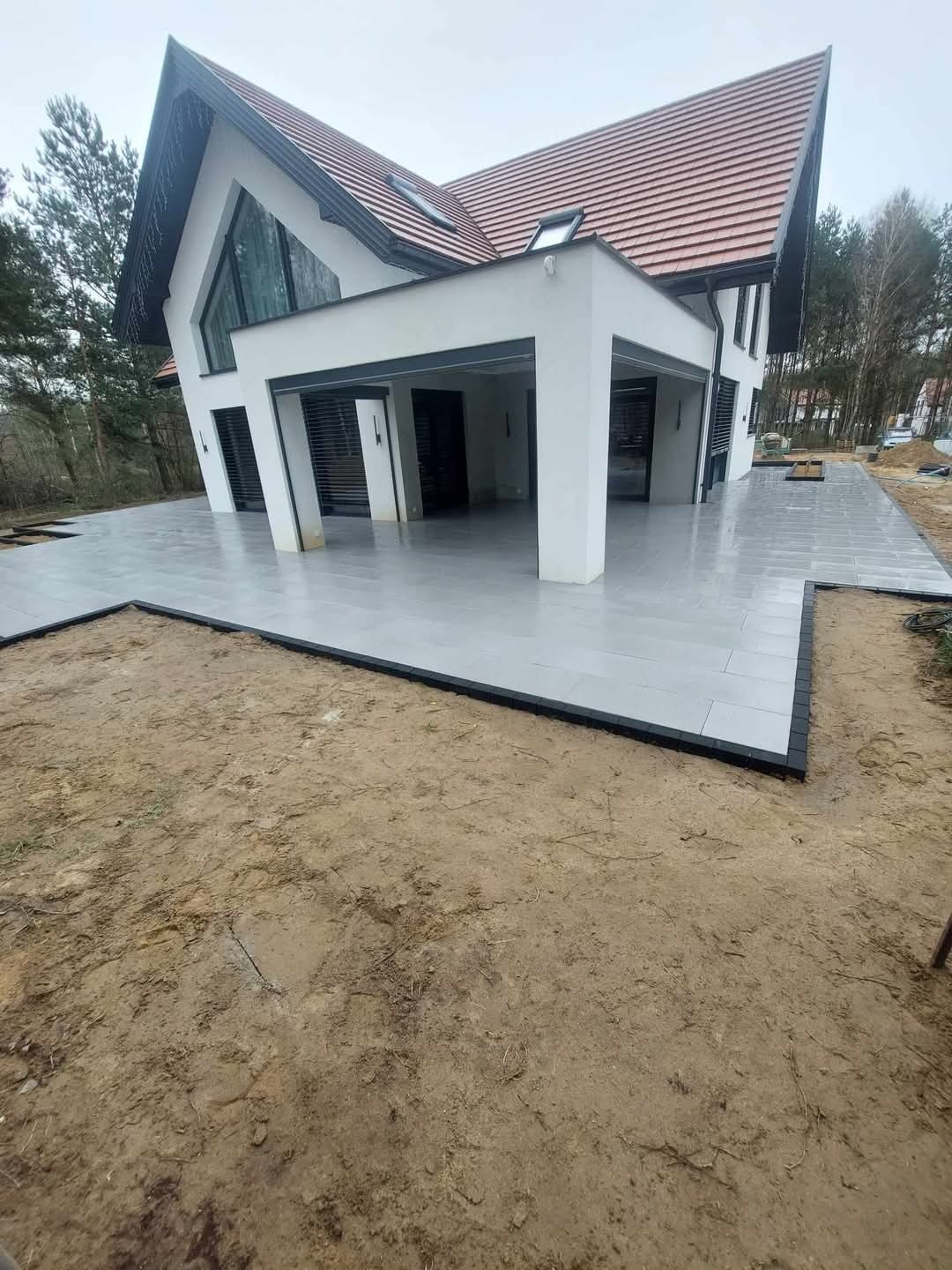 Nowoczesny taras z szarych płyt, obrzeża z ciemnej kostki, dom z dużym oknem trójkątnym w tle. Realizacja w Morągu.