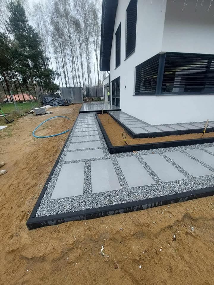 Nowoczesna ścieżka z płyt betonowych i grysu, obrzeża z czarnej kostki, prowadząca wzdłuż białego budynku z żaluzjami. Widok z poziomu gruntu.