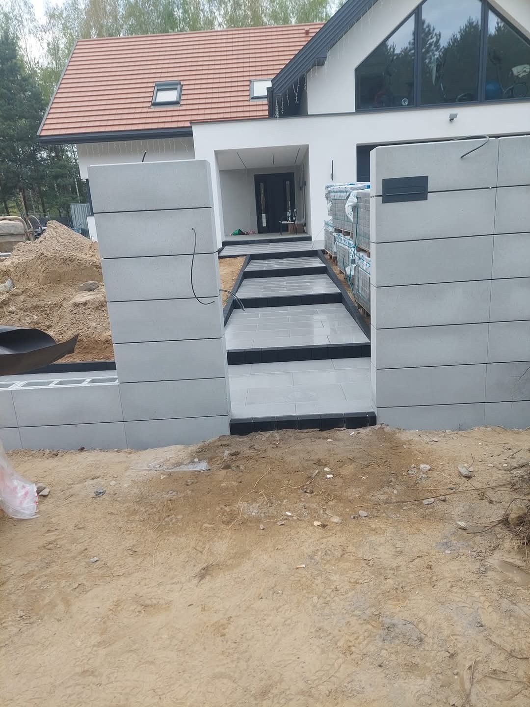 Nowoczesne podejście do domu z szarej kostki, obrzeża z czarnego granitu. Widoczne palety z kostką. Częściowo wykonane ogrodzenie z betonowych bloczków.