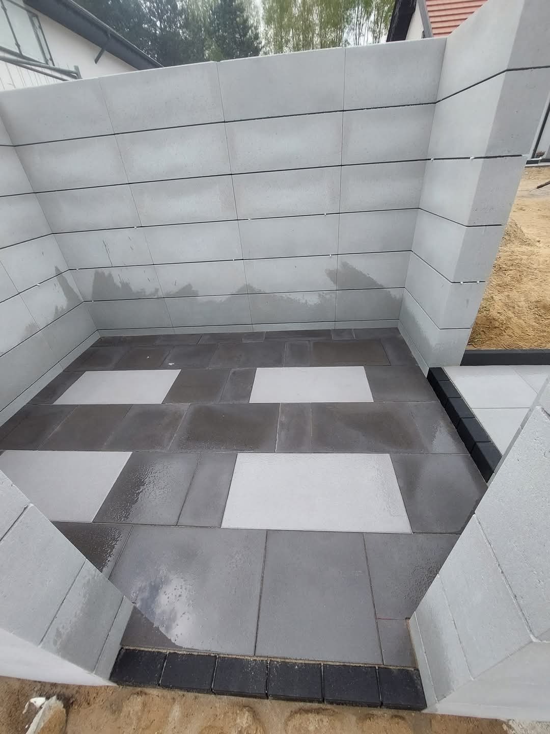 Nowoczesna aranżacja tarasu z szarej kostki brukowej i betonowych ścianek. Kontrastujące, jasne płyty dodają unikalnego charakteru. Widoczne detale wykończenia.