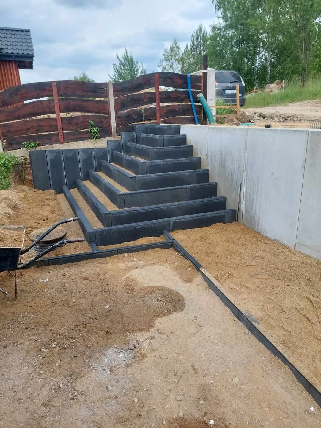 Schody z kostki brukowej prowadzące w górę, obok betonowa ściana i drewniane ogrodzenie. Widoczny piasek i elementy konstrukcyjne.