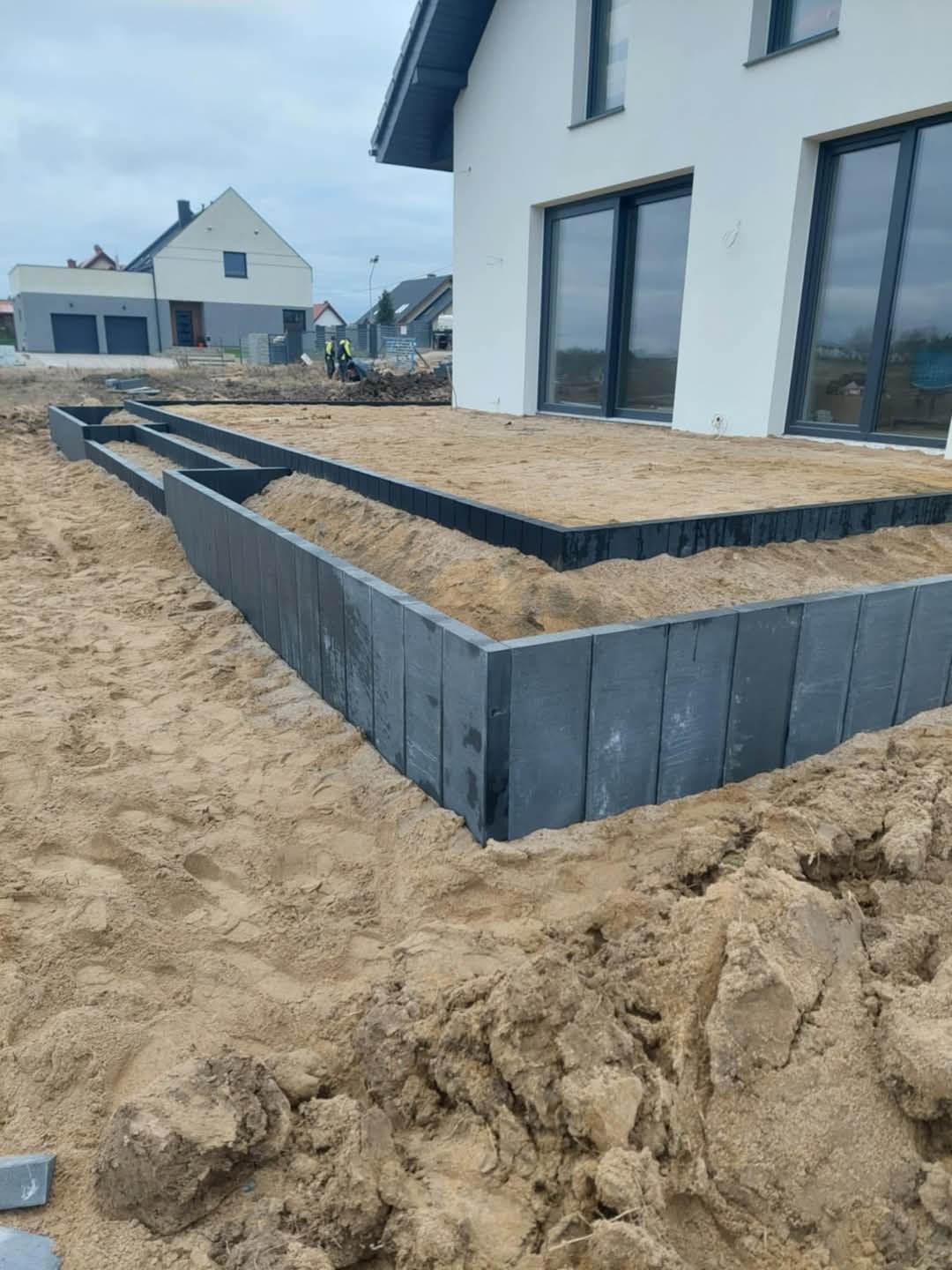 Przygotowanie podłoża pod taras z obrzeżami z betonowych płyt. W tle dom z dużymi oknami i ekipa budowlana. Piasek, ziemia i elementy konstrukcyjne.