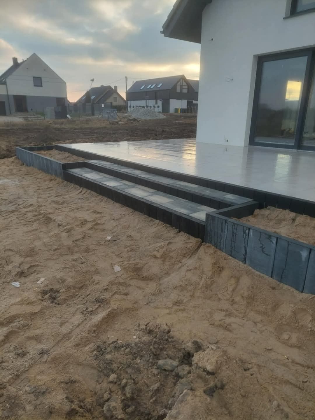 Nowoczesny taras z kostki brukowej w odcieniach szarości, z wyraźnie zaznaczonymi schodami i obrzeżami. W tle dom jednorodzinny i przygotowany teren pod dalsze prace.