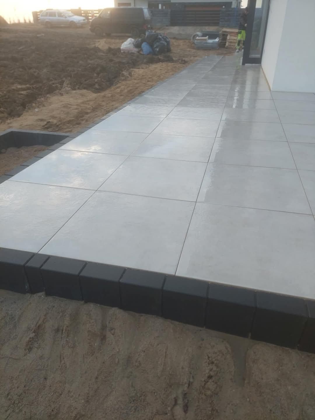 Ułożona jasnoszara kostka brukowa na tarasie z czarnym obrzeżem, widok z bliska na świeżo wykonaną nawierzchnię. W tle ziemia i budynek.