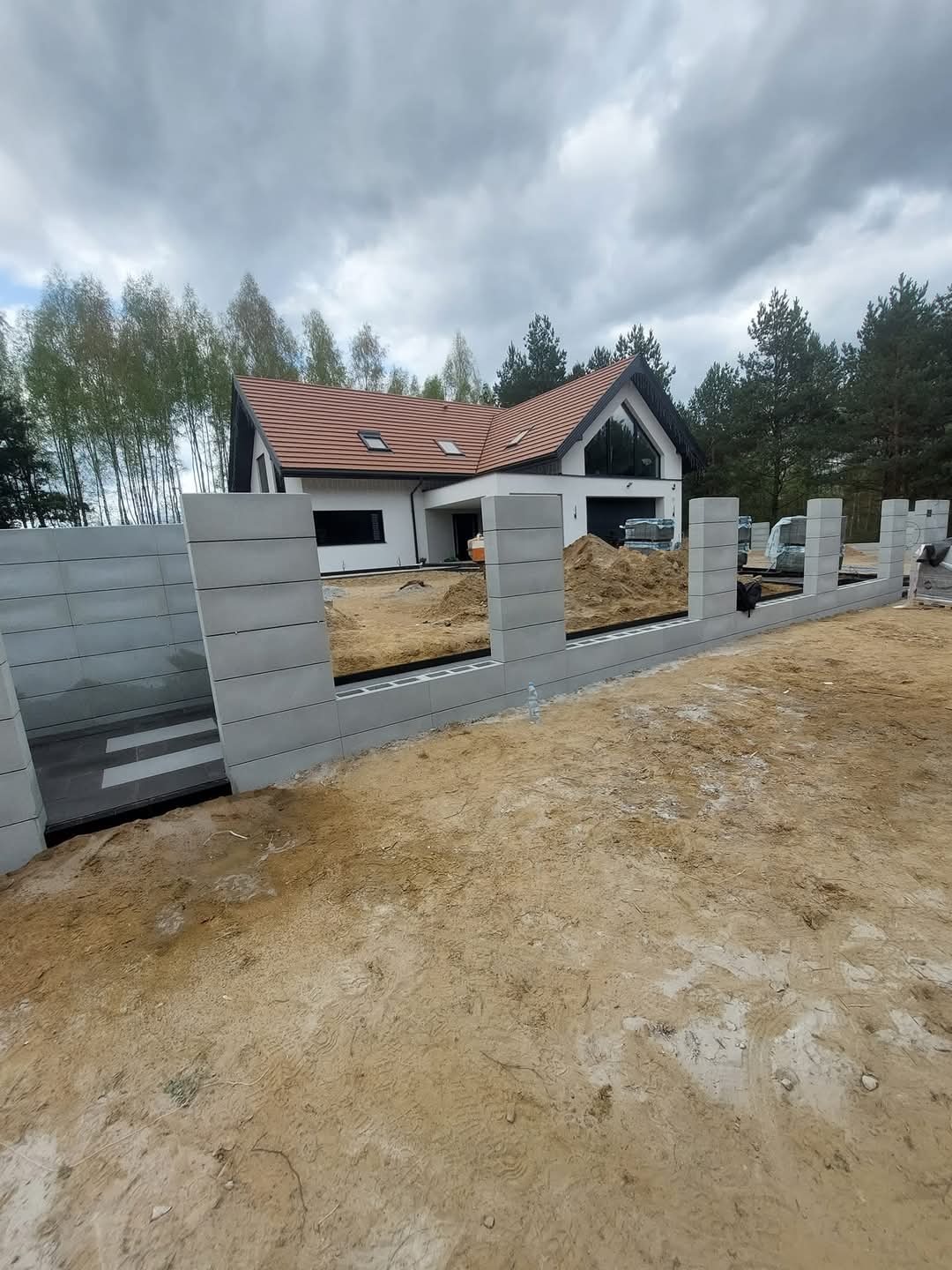 Wznoszenie ogrodzenia z szarych, betonowych bloczków modułowych na posesji z nowoczesnym domem. Widoczne elementy konstrukcyjne i przygotowany teren.