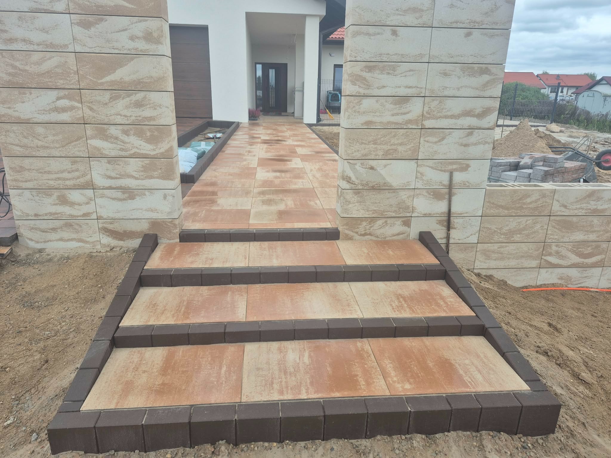 Schody i chodnik z kostki brukowej w odcieniach beżu i brązu prowadzące do domu. Widoczne elementy wykończeniowe i przygotowany teren.