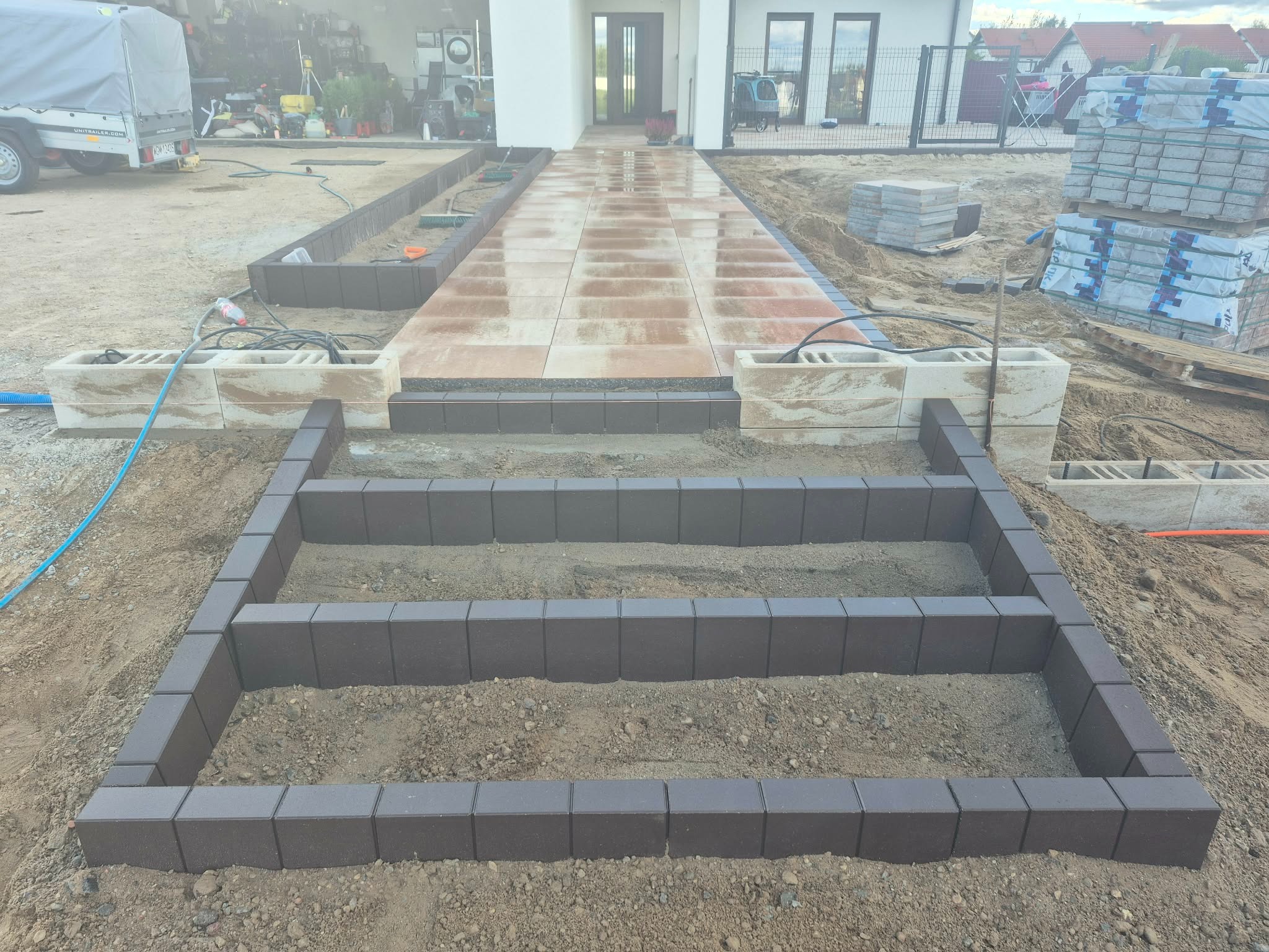 Budowa schodów wejściowych z brązowej kostki brukowej, widok z góry na plac budowy przed domem z ułożonym chodnikiem z jasnych płyt. Częściowo ułożona kostka.
