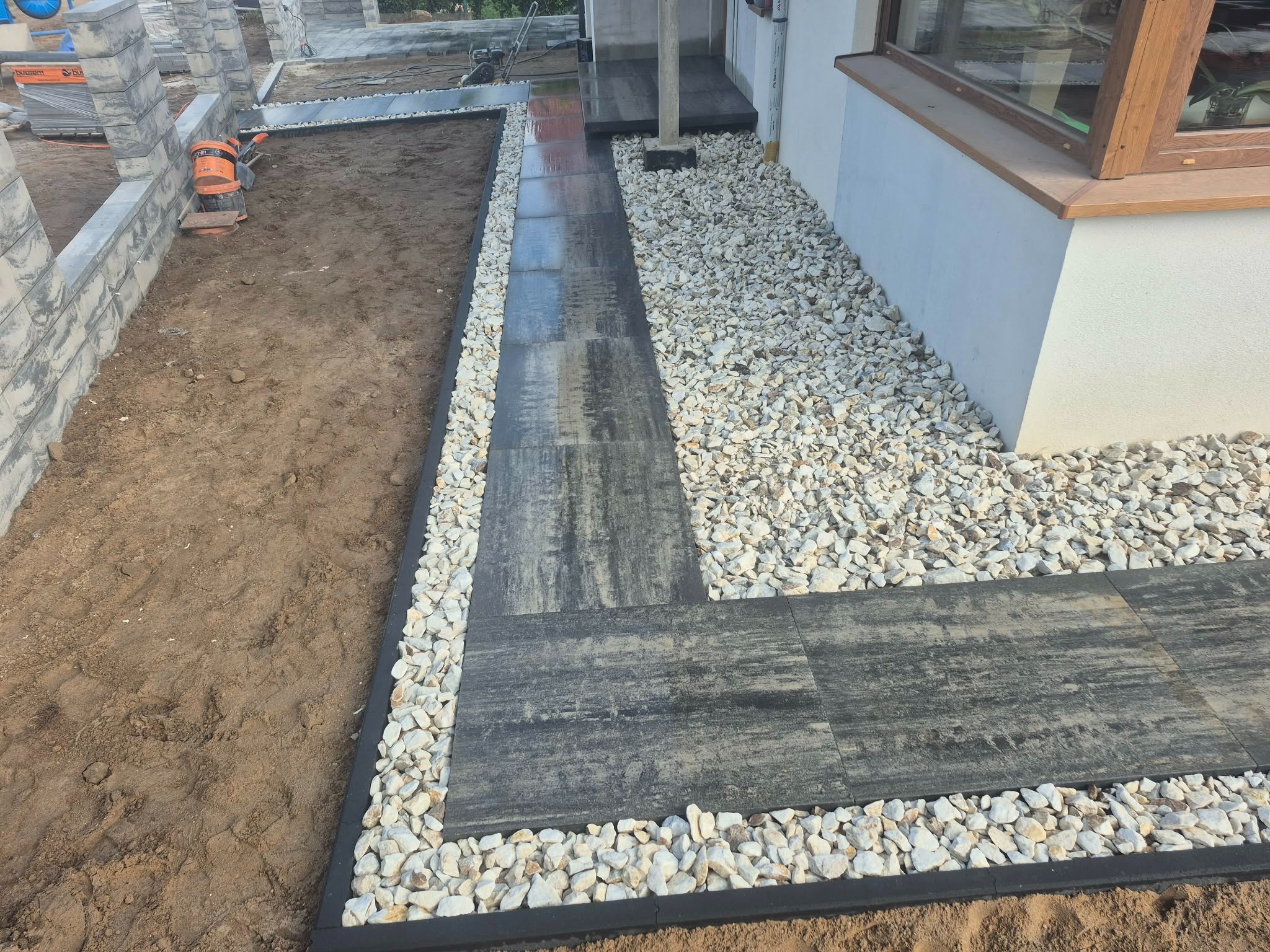 Nowoczesna ścieżka z szarych płyt, obłożona białym kamieniem, prowadząca wzdłuż budynku. Widoczne elementy ogrodzenia w trakcie budowy i przygotowany teren.