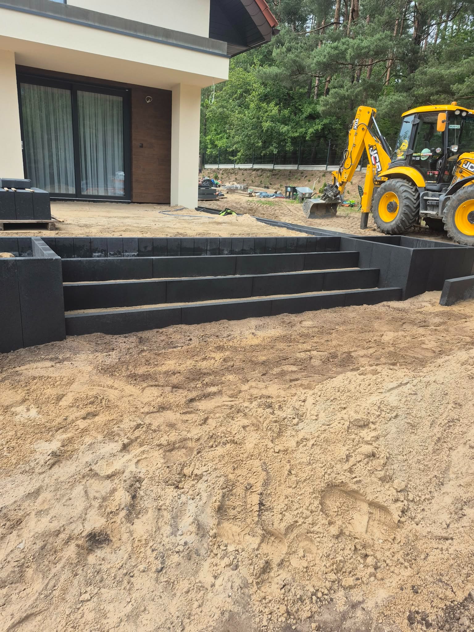 Schody z kostki brukowej w ogrodzie, z żółtą koparką w tle. Widoczne świeże ślady opon na piasku. Nowoczesny dom z dużym oknem przesuwnym.