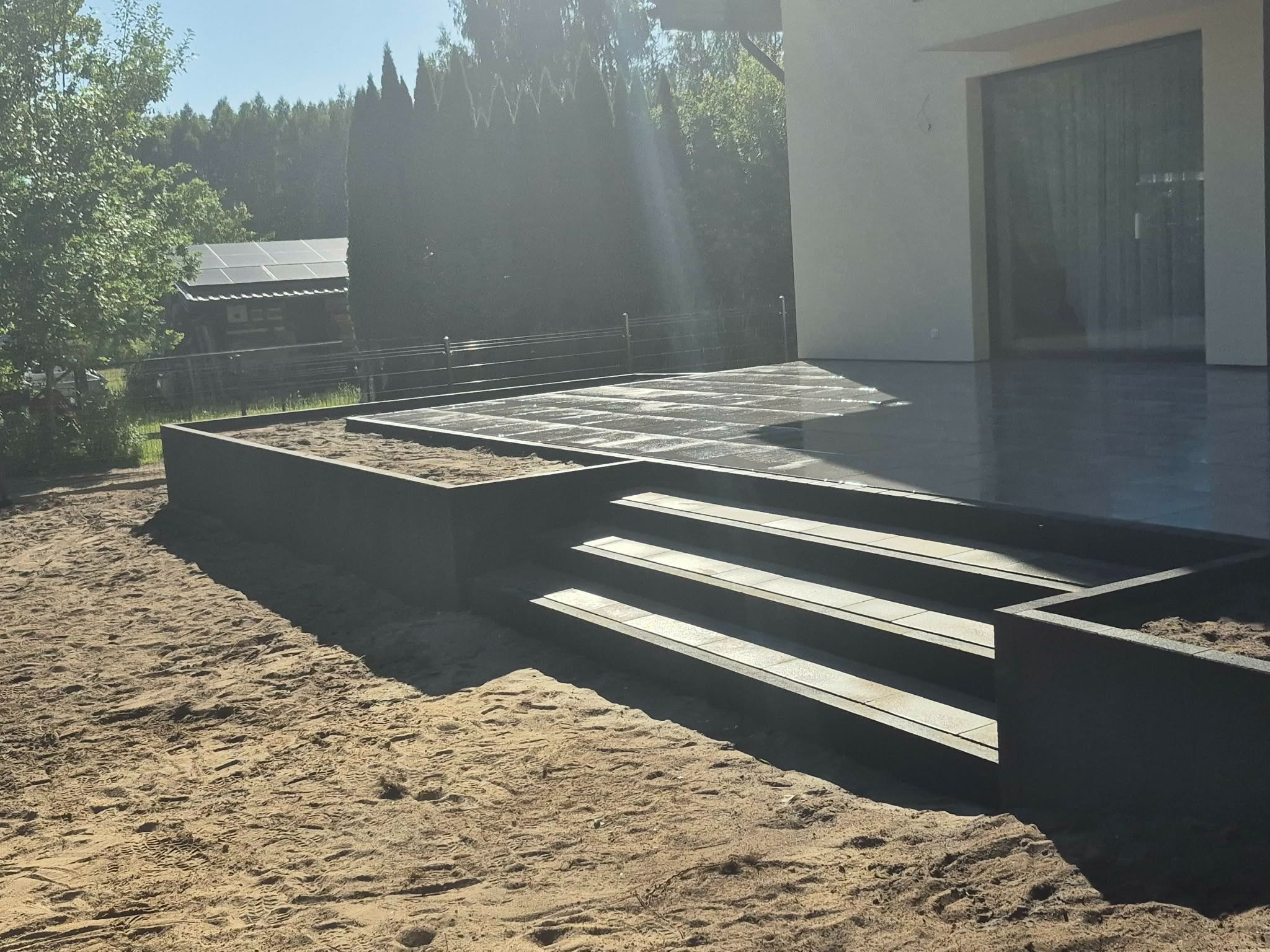 Nowoczesne schody z kostki brukowej prowadzące na taras, otoczone piaskiem. Widoczny fragment domu z dużym oknem i zieleń w tle.