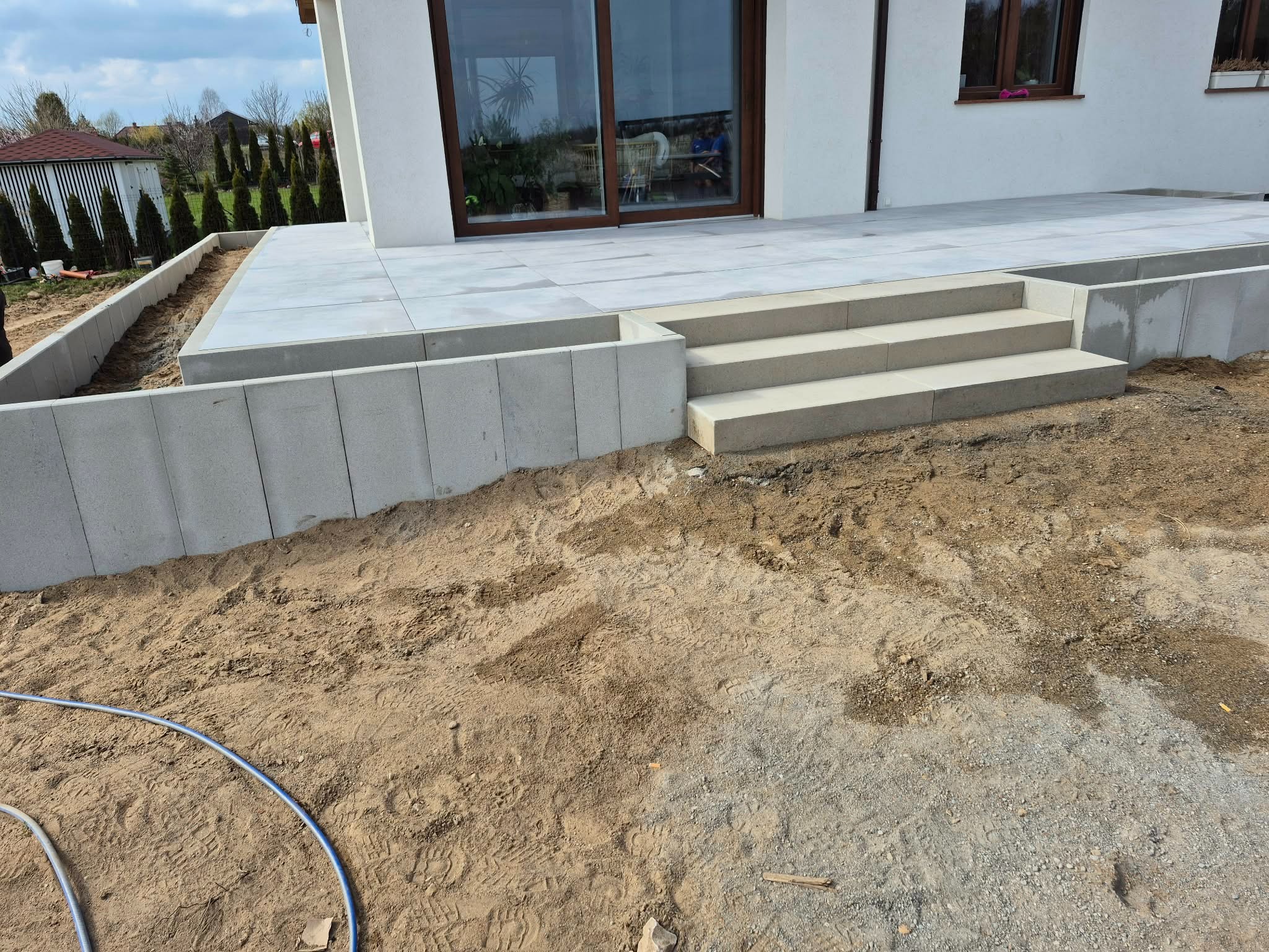 Nowoczesny taras z jasnoszarej kostki brukowej i schodami, otoczony betonowymi murkami oporowymi na piaszczystym podłożu. Widok na dom z dużymi oknami.