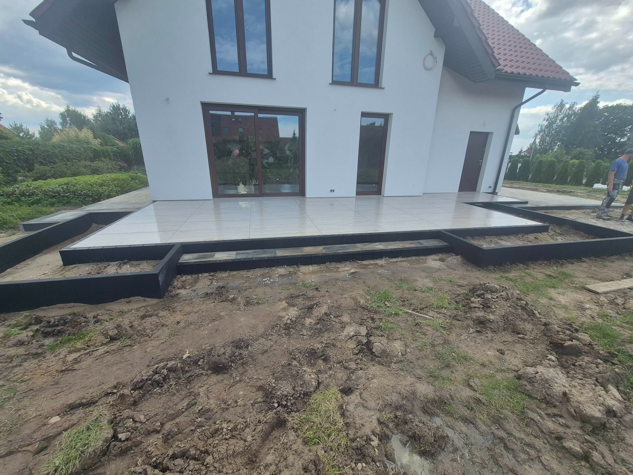 Wykończony taras z jasnych płytek przy białym domu z brązowymi oknami. Widoczne czarne obramowanie tarasu i przygotowany teren wokół. Prace w toku.