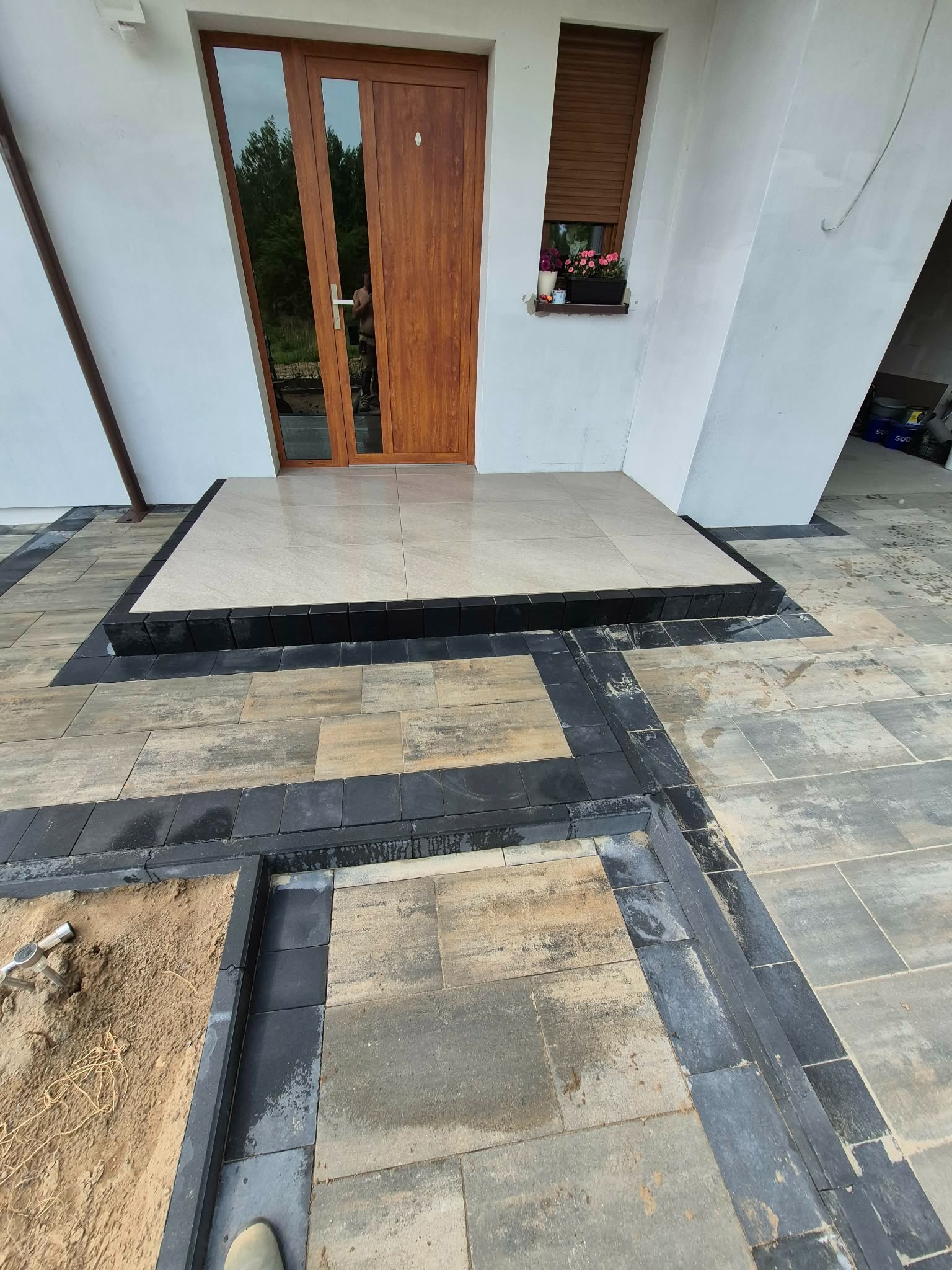 Wykończone wejście do domu z jasnych płyt i ciemnej kostki. Fragment niedokończonej powierzchni z piaskiem i narzędziem. Widoczne drzwi wejściowe i okno z kwiatami.