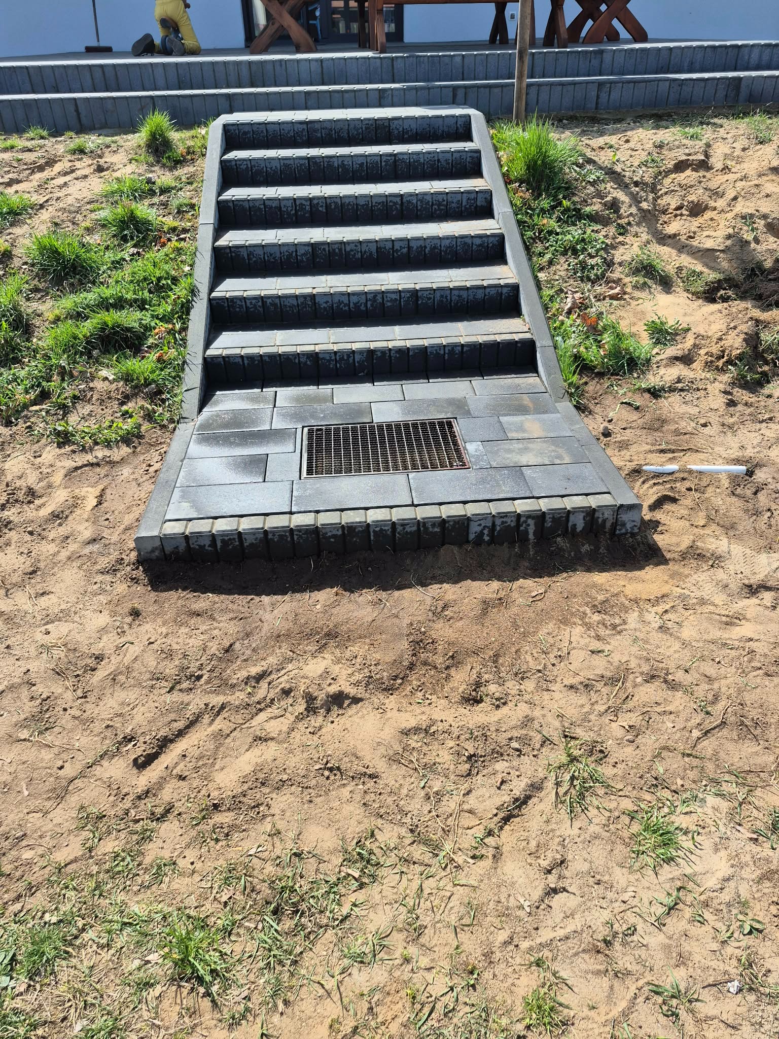 Schody z kostki brukowej prowadzące w górę zbocza, z kratką odpływową na dole. Widoczne ślady prac ziemnych i trawa. Ujęcie z dołu, podkreślające solidność konstrukcji.