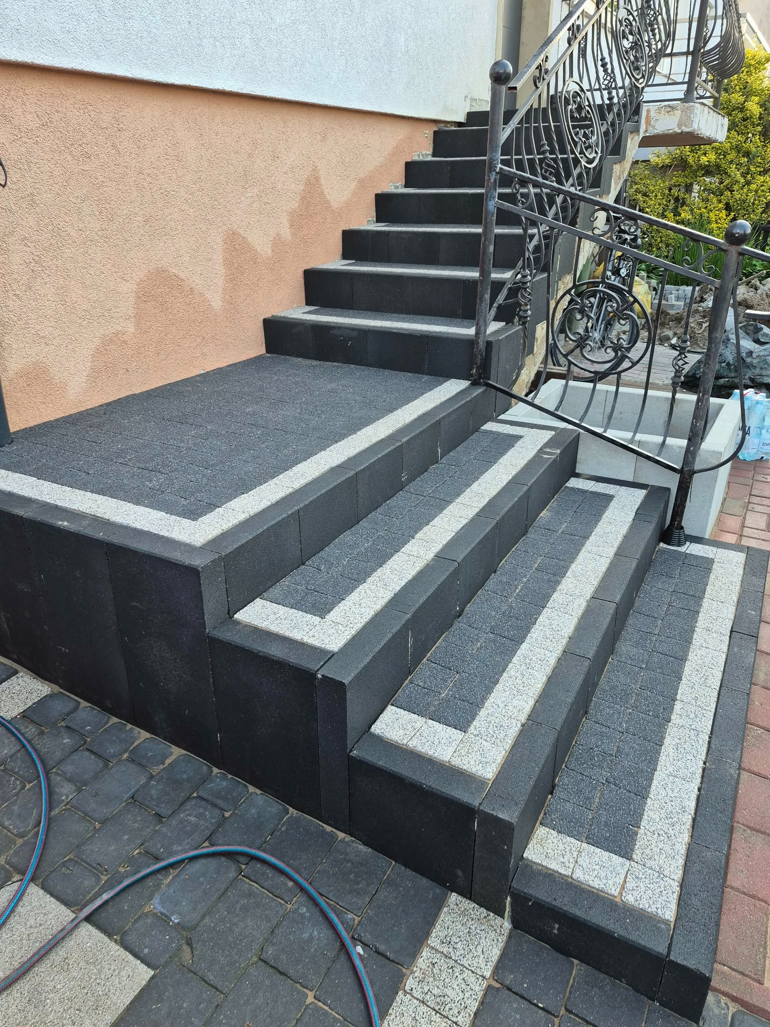 Zewnętrzne schody z kostki brukowej w odcieniach szarości, obramowane jaśniejszą kostką. Metalowa balustrada z ozdobnymi elementami. Częściowo widoczny wąż ogrodowy.