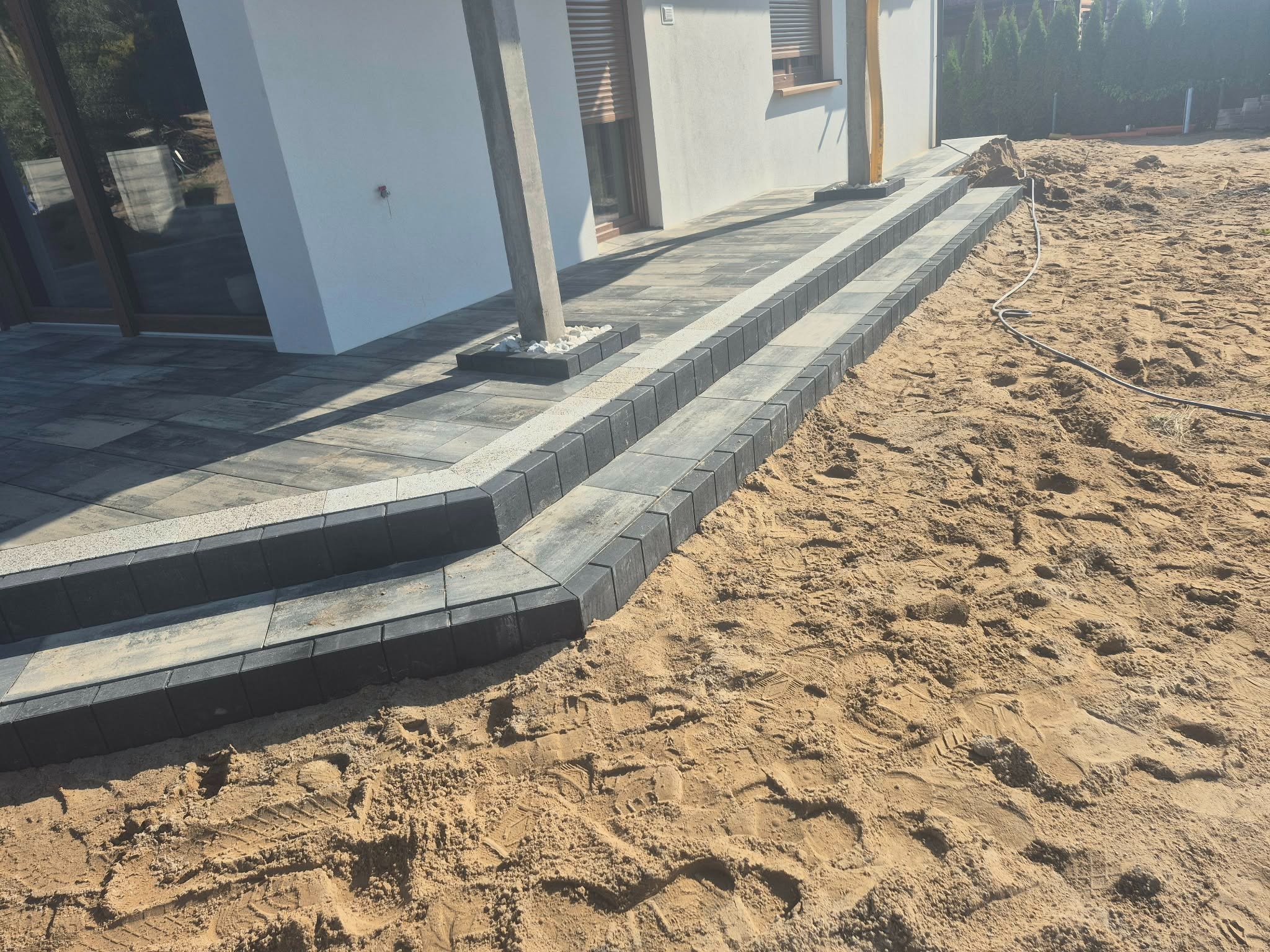 Nowoczesne schody z kostki brukowej prowadzące do domu, z widocznym przygotowanym terenem pod dalsze prace. Jasne, naturalne światło.