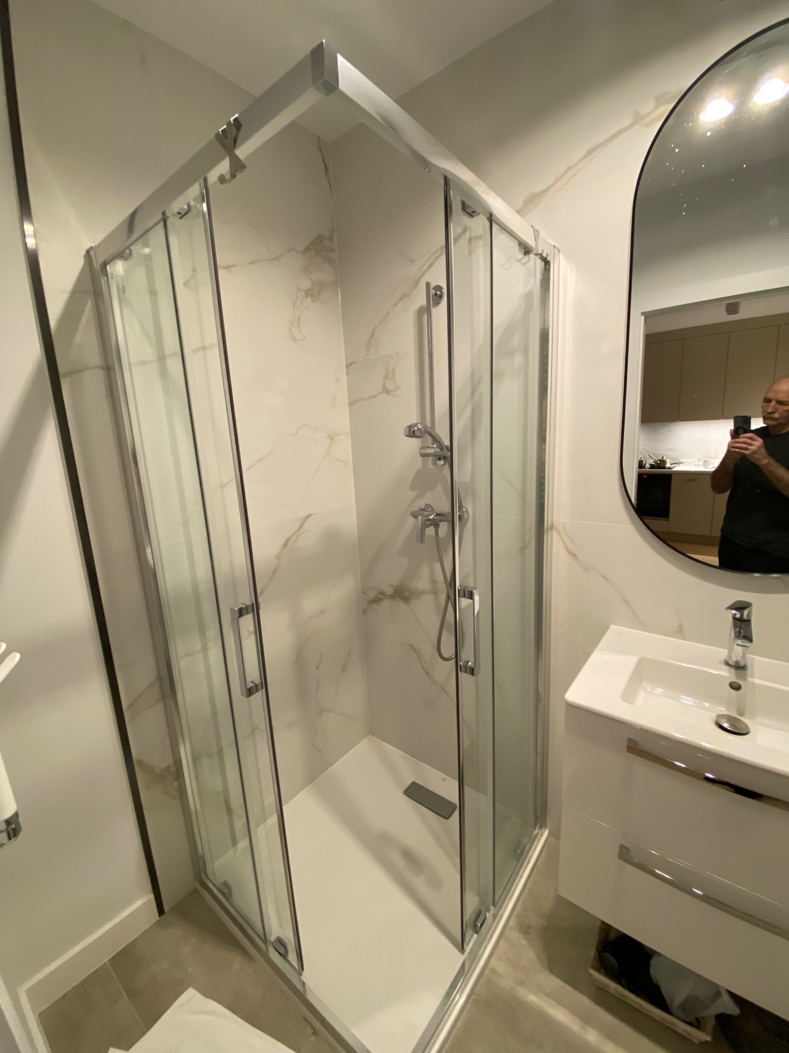 Nowoczesna kabina prysznicowa z armaturą w wykończonej łazience. W lustrze odbija się osoba robiąca zdjęcie. Białe marmurowe płytki na ścianach.