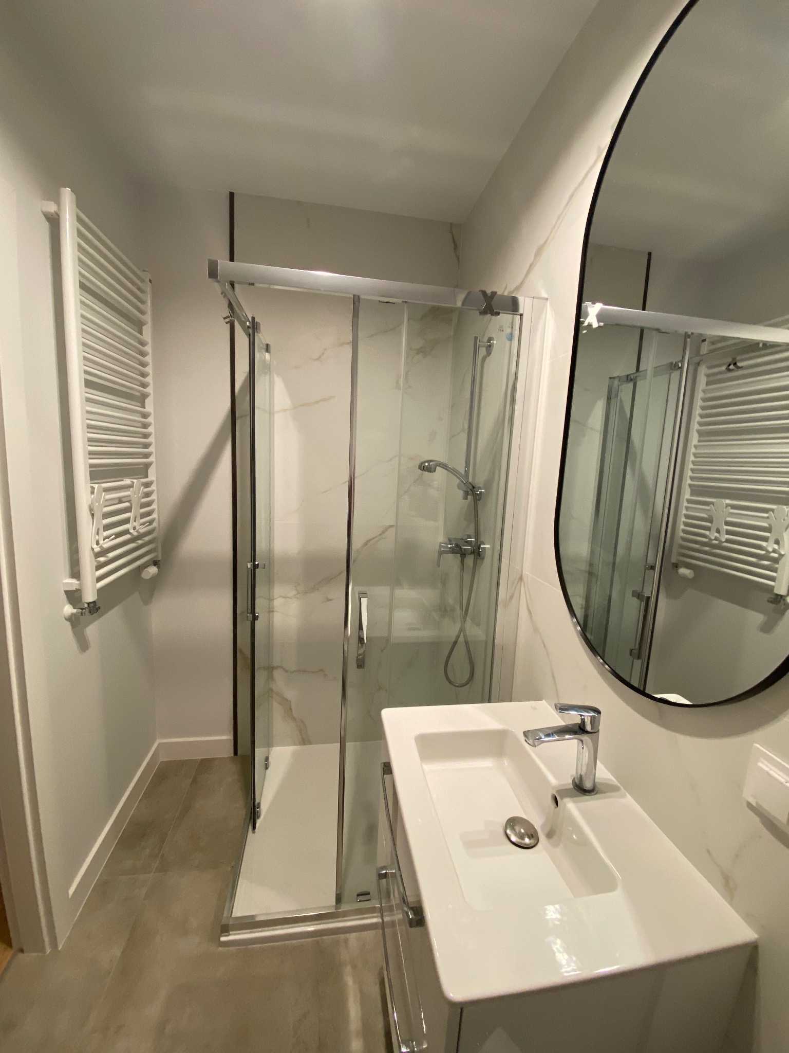 Nowoczesna łazienka z kabiną prysznicową, umywalką i grzejnikiem drabinkowym. Marmurowe płytki na ścianach, owalne lustro. Minimalistyczny design, jasne kolory.