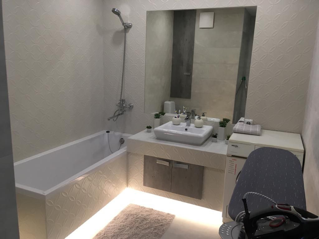 Jasna łazienka z wanną, umywalką i podświetleniem pod szafką. Na blacie umywalki rośliny w doniczkach. Na pralce deska do prasowania. Ściany z wzorem.