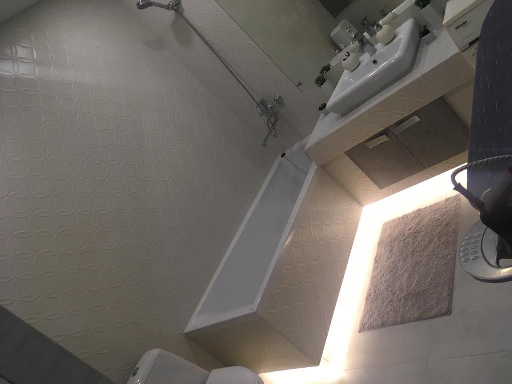 Nowoczesna łazienka z białą armaturą, dekoracyjnymi płytkami ściennymi i subtelnym oświetleniem LED. Umywalka nablatowa, minimalistyczna bateria i lustro.