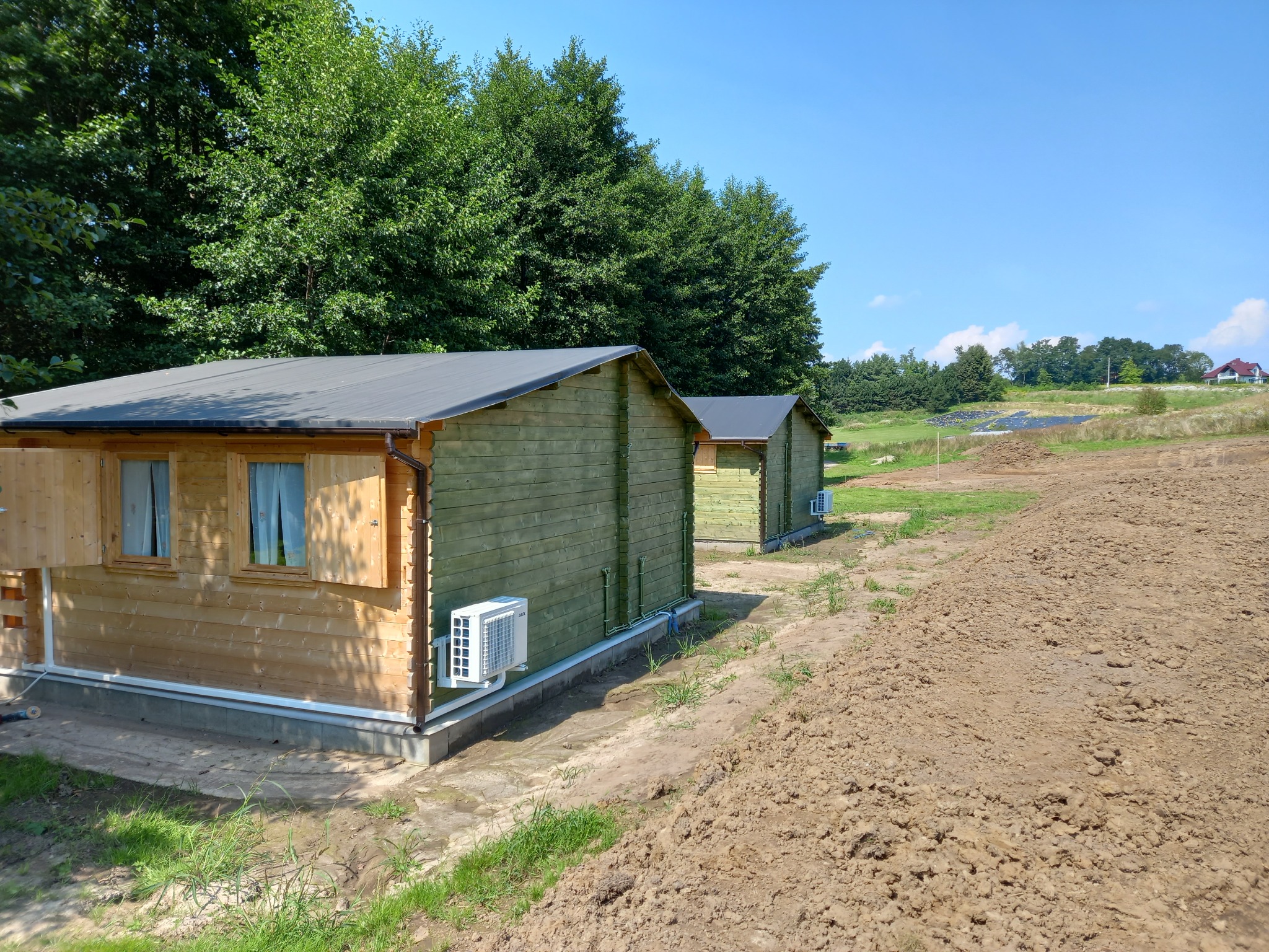 Dwa drewniane domki z klimatyzacją na zewnątrz, na tle zieleni i błękitnego nieba. Widoczne panele słoneczne w oddali. Ujęcie w słoneczny dzień.