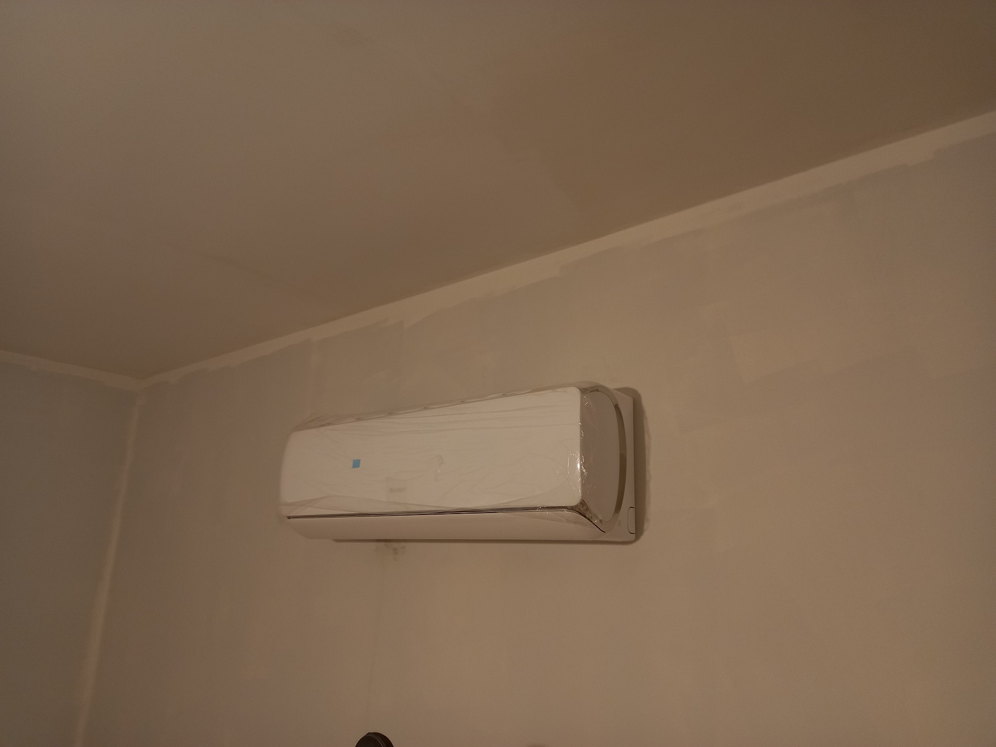 Nowy, biały klimatyzator ścienny, jeszcze w folii ochronnej, zamontowany w rogu pokoju o beżowych ścianach. Widoczne niedomalowania na ścianie.