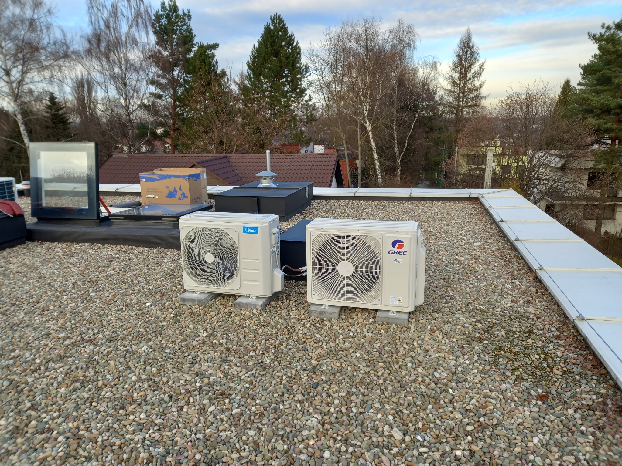 Dwa klimatyzatory Midea i Gree na żwirowym dachu budynku z widokiem na drzewa i inne budynki. Widoczne elementy wentylacji i okno dachowe.