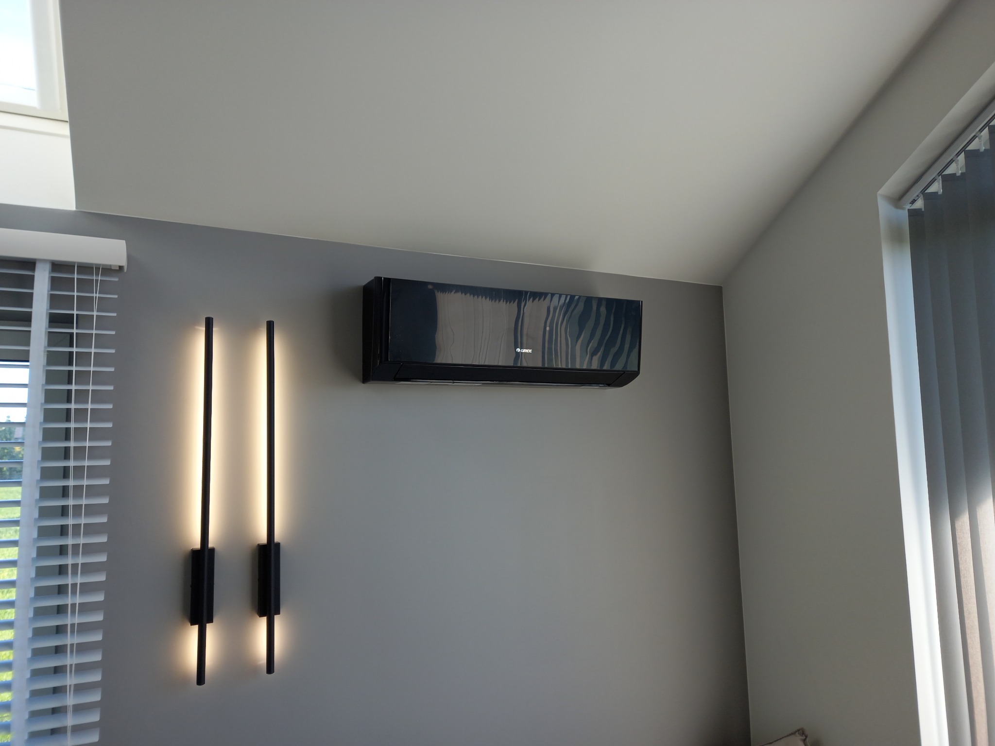 Czarny klimatyzator Gree zamontowany na szarej ścianie w pokoju, obok pionowe lampy LED i okno z żaluzjami. Nowoczesny design, minimalistyczne wnętrze.