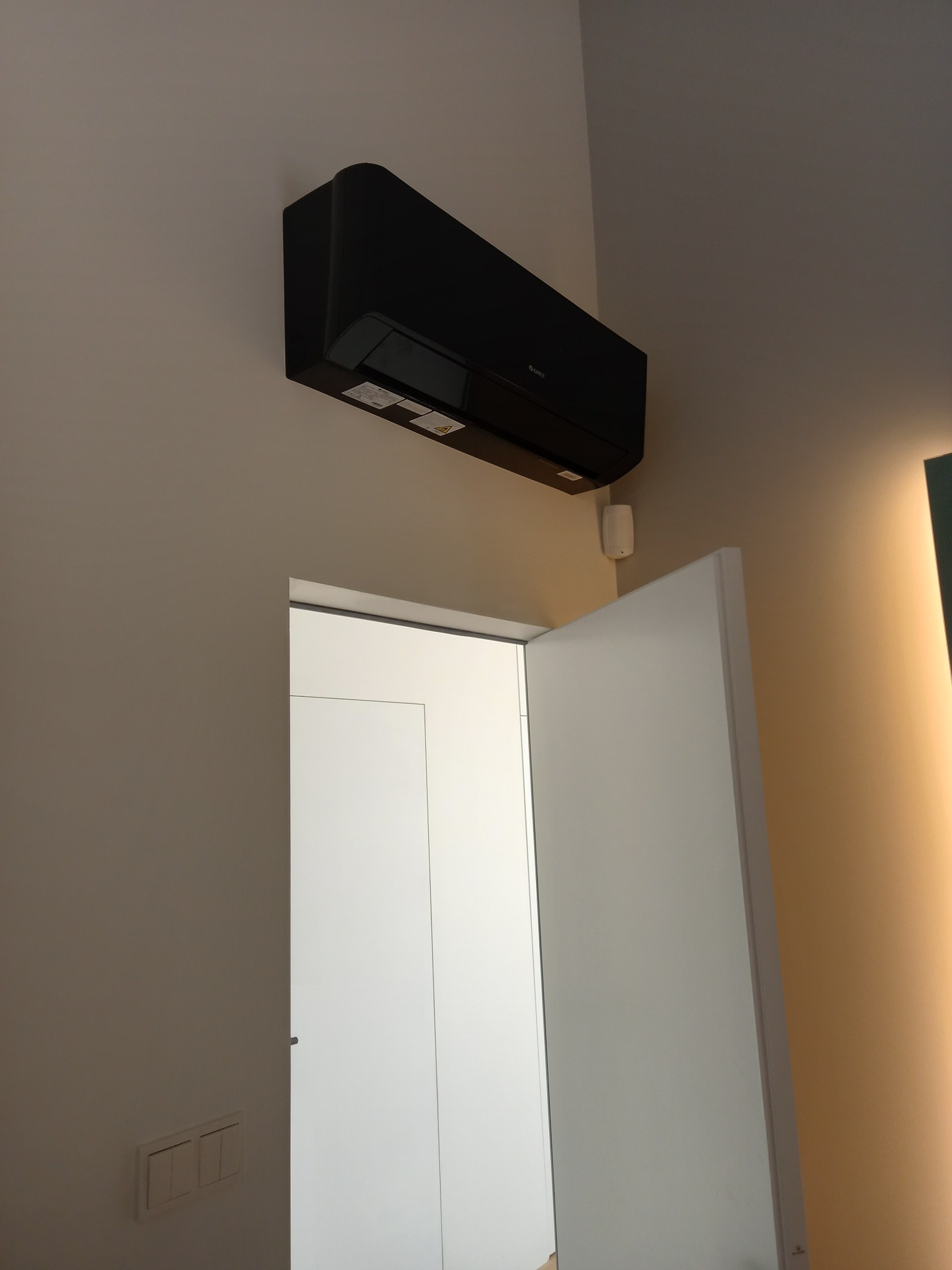 Czarny klimatyzator ścienny w nowoczesnym wnętrzu, widoczny nad otwartymi białymi drzwiami. Minimalistyczny design, jasne ściany, oświetlenie akcentujące.
