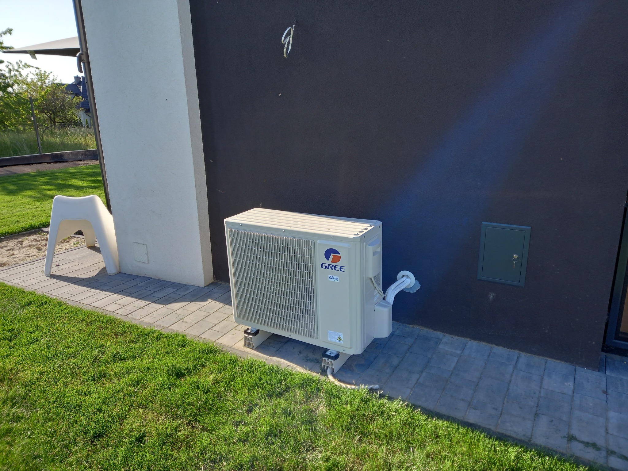 Kompaktowa jednostka zewnętrzna klimatyzacji Gree, zamontowana na kostce brukowej przy ciemnej ścianie budynku z zielonym trawnikiem. Widoczne przewody i skrzynka rewizyjna.