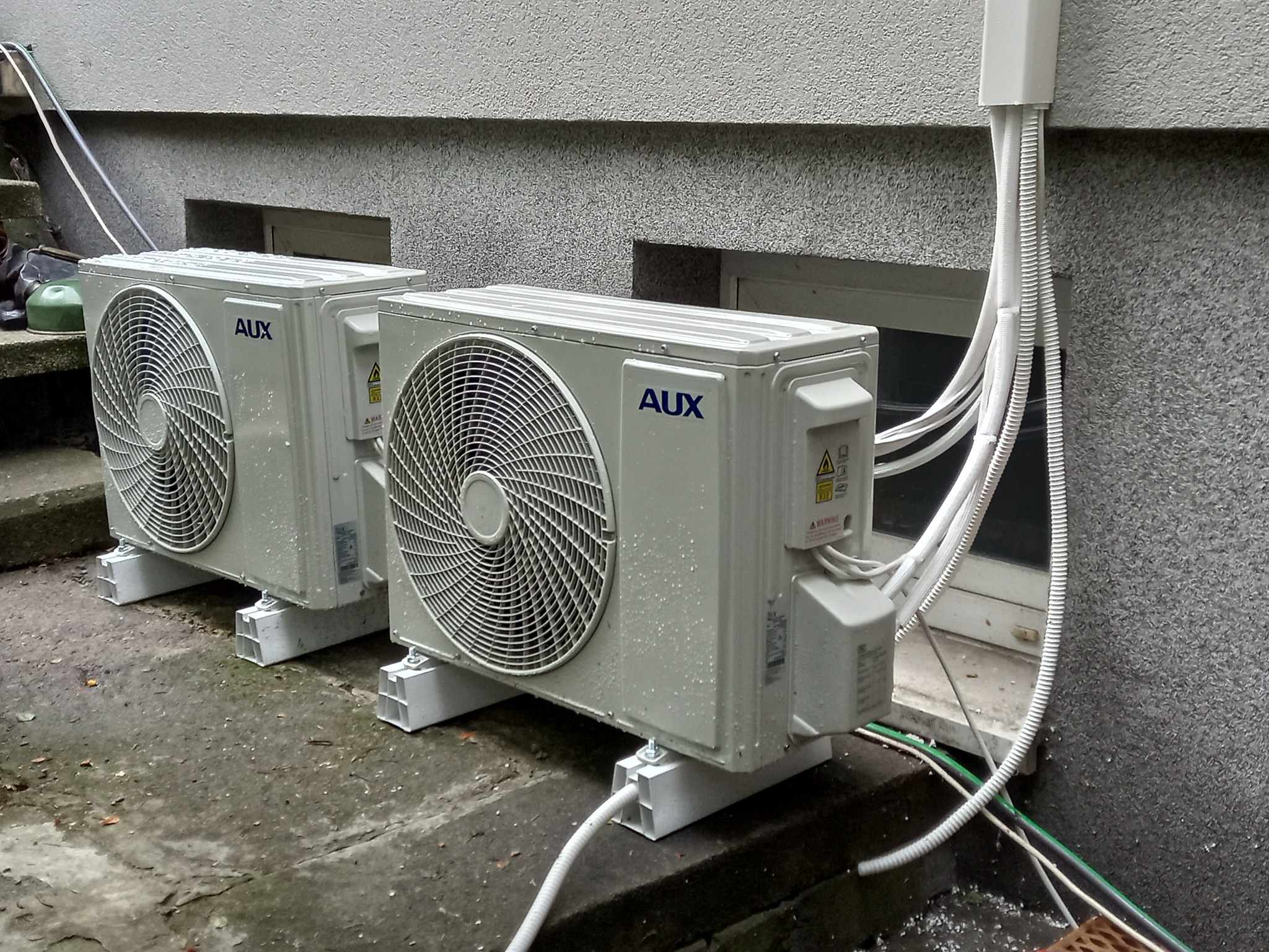 Dwa klimatyzatory AUX zamontowane na zewnątrz budynku, z widocznymi przewodami i instalacją elektryczną. Ściana budynku z szarą, chropowatą fakturą. Widoczne krople wody na urządzeniach.