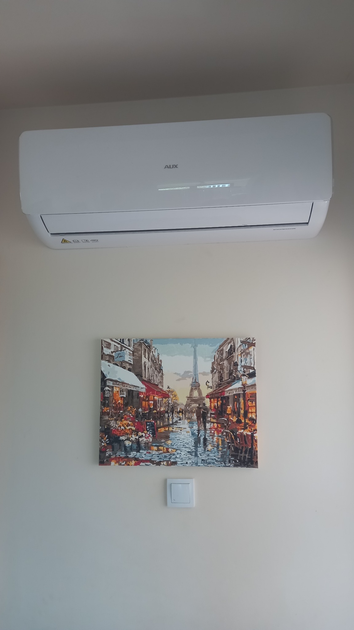 Biały klimatyzator AUX zamontowany na ścianie nad obrazem z wieżą Eiffla. Widoczny włącznik światła. Minimalistyczny design wnętrza.