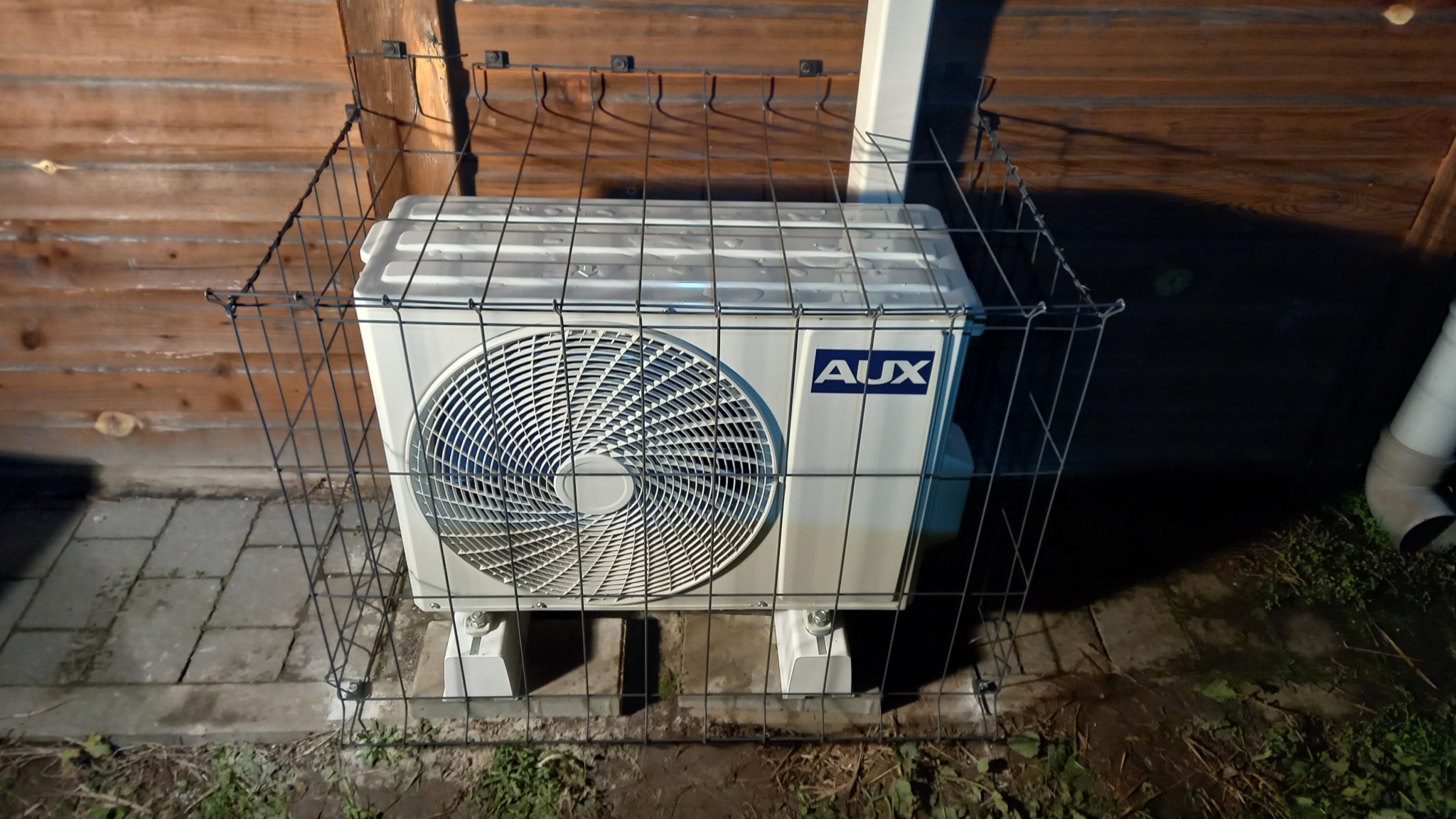 Biała jednostka zewnętrzna klimatyzacji AUX, zabezpieczona metalową klatką, stoi na betonowych płytach przy drewnianym ogrodzeniu. Widoczne elementy instalacji.