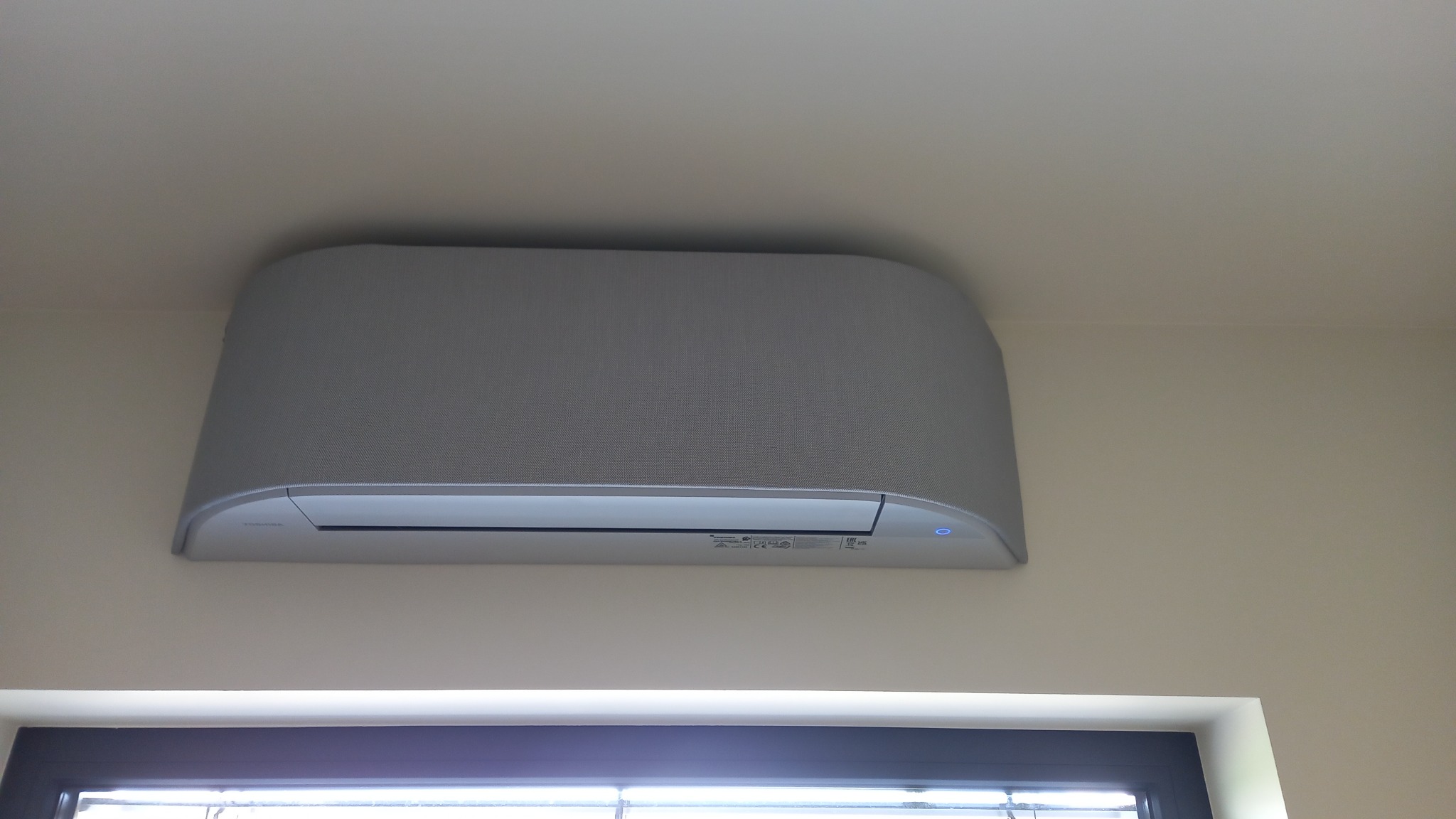 Szary klimatyzator Toshiba zamontowany na ścianie nad oknem, widoczny z dołu, z delikatnym oświetleniem i fakturą obudowy.