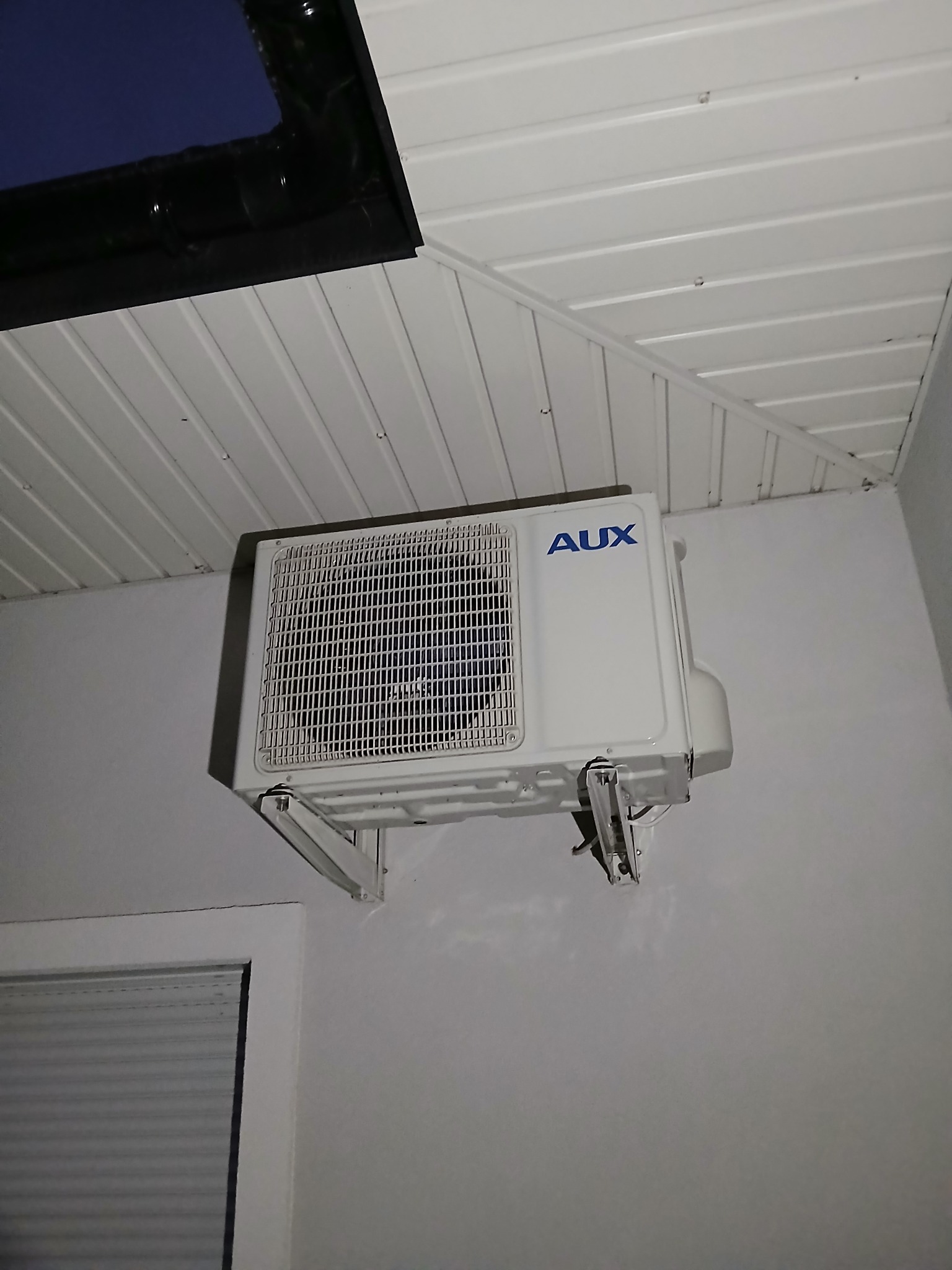 Biała jednostka zewnętrzna klimatyzacji AUX zamontowana na ścianie wewnątrz pomieszczenia z widocznym oknem i skośnym sufitem z boazerii.