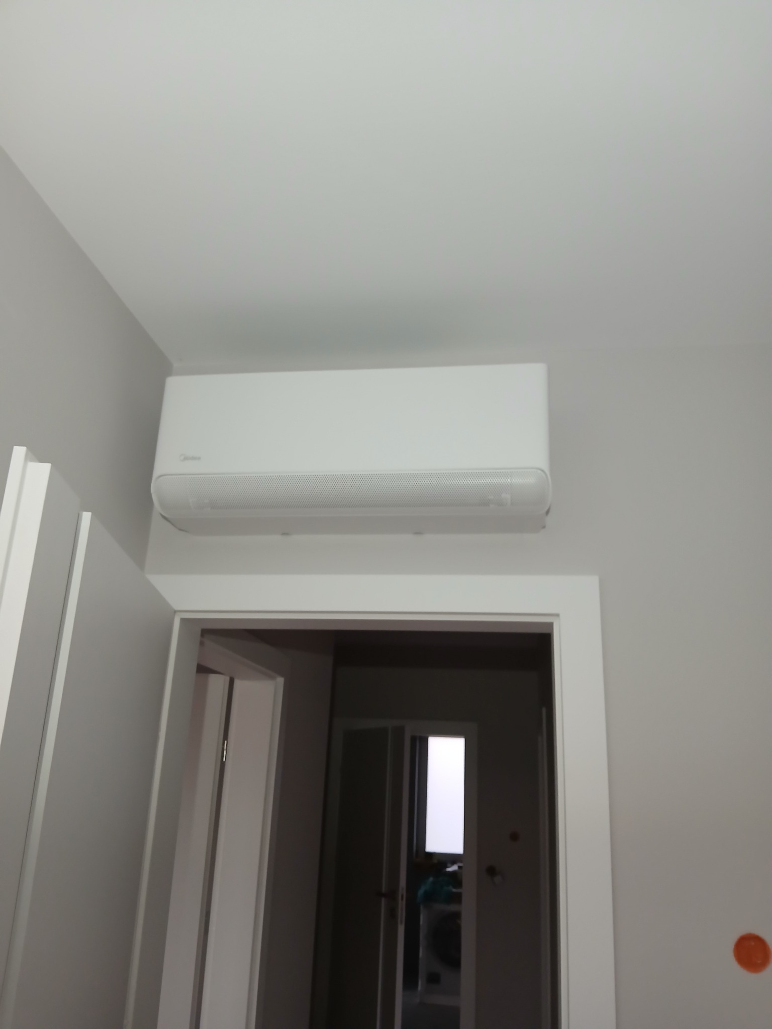 Biały klimatyzator Midea zamontowany nad drzwiami w jasnym pomieszczeniu. Widok z dołu, neutralne oświetlenie, minimalistyczny design wnętrza.