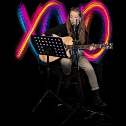 FotoBokeh - Kobieta z długimi włosami śpiewa do mikrofonu, grając na gitarze Yamaha. W tle kolorowe, rozmyte światła tworzą efektowny wizualny akcent. Scena koncertowa.