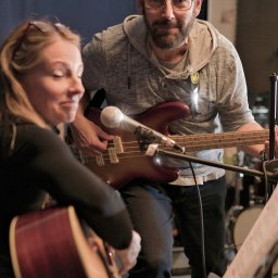 FotoBokeh - Zespół muzyczny w trakcie próby: kobieta gra na gitarze akustycznej, mężczyzna na gitarze basowej, mikrofon na statywie, nuty, perkusja w tle. Kameralna atmosfera.