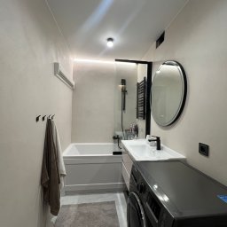 CimentArt Polska - Nowoczesna łazienka w beżowych tonacjach z wanną, umywalką, okrągłym lustrem i pralką. Minimalistyczny design, oświetlenie LED i szary dywanik.