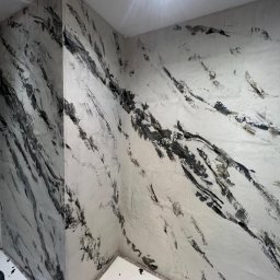 CimentArt Polska - Ściana w łazience wykończona mikrocementem w odcieniach bieli i szarości, imitująca marmurkowe wzory. Widoczny narożnik i fragment podłogi z kratką ściekową.