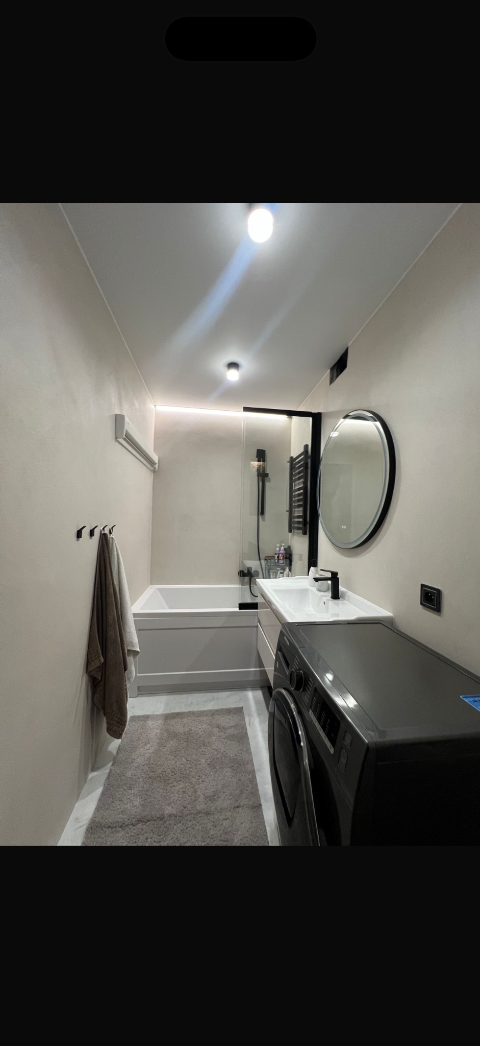 Nowoczesna łazienka w beżowych tonacjach z wanną, umywalką, okrągłym lustrem i pralką. Minimalistyczny design, oświetlenie LED i szary dywanik.