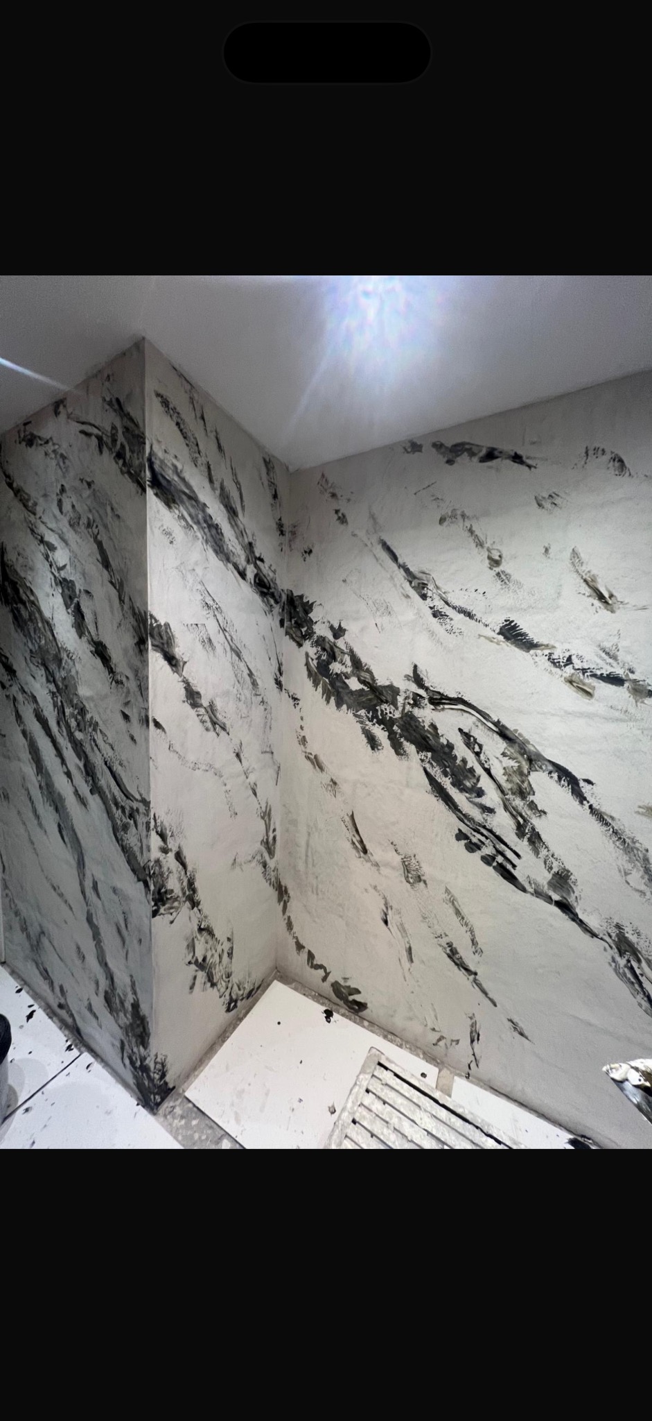 Ściana w łazience wykończona mikrocementem w odcieniach bieli i szarości, imitująca marmurkowe wzory. Widoczny narożnik i fragment podłogi z kratką ściekową.