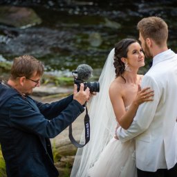 Studio Kadr Łukasz Michalik - Kamerzysta filmuje parę młodą na tle strumienia. Ujęcie z boku, widoczny mikrofon na kamerze Lumix S5. Panna młoda w sukni ślubnej, pan młody w białym smokingu.