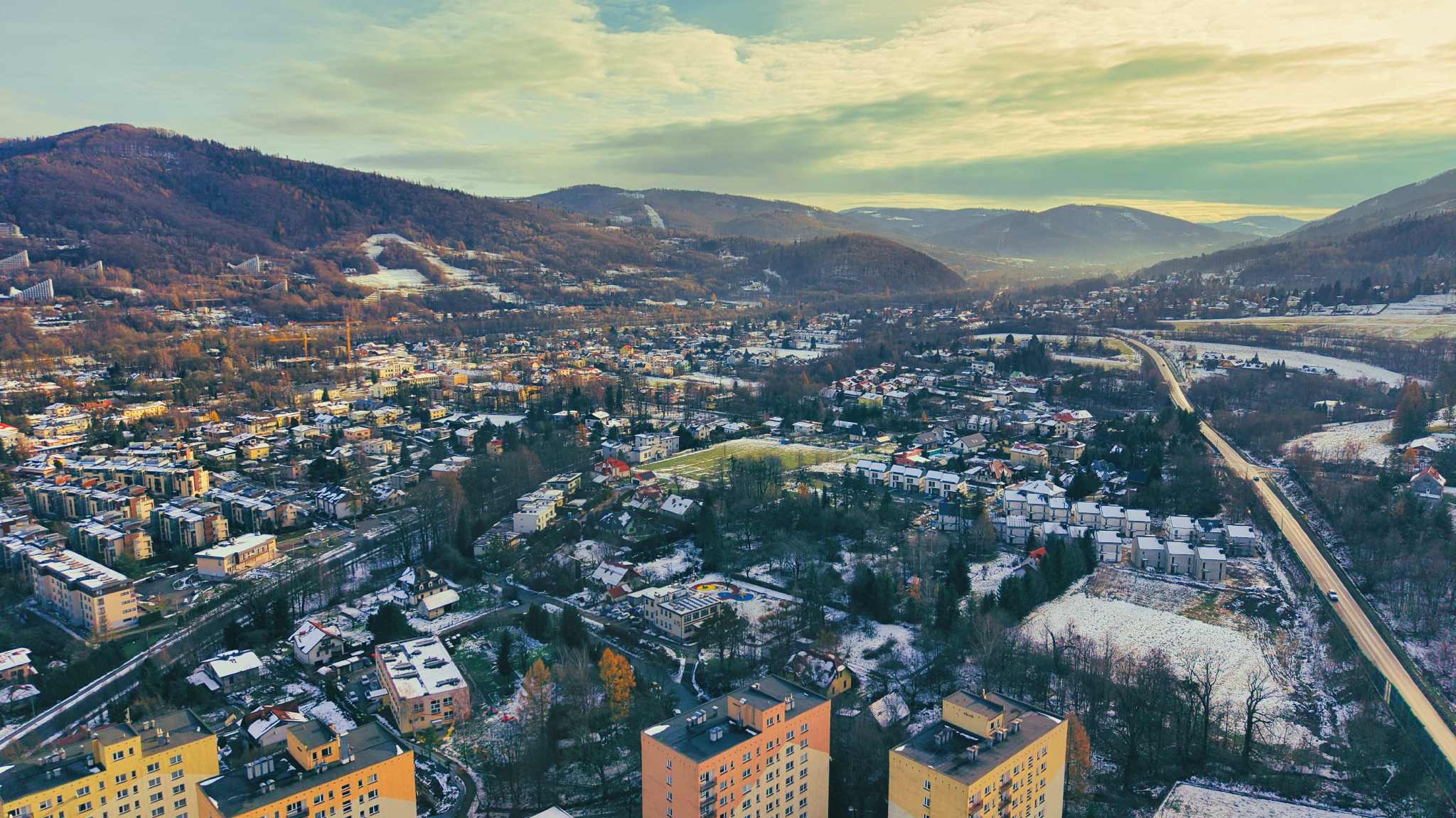 Zimowy krajobraz miejski z lotu ptaka: budynki mieszkalne, góry w tle, lekko ośnieżone dachy i drzewa, ciepłe światło zachodzącego słońca.