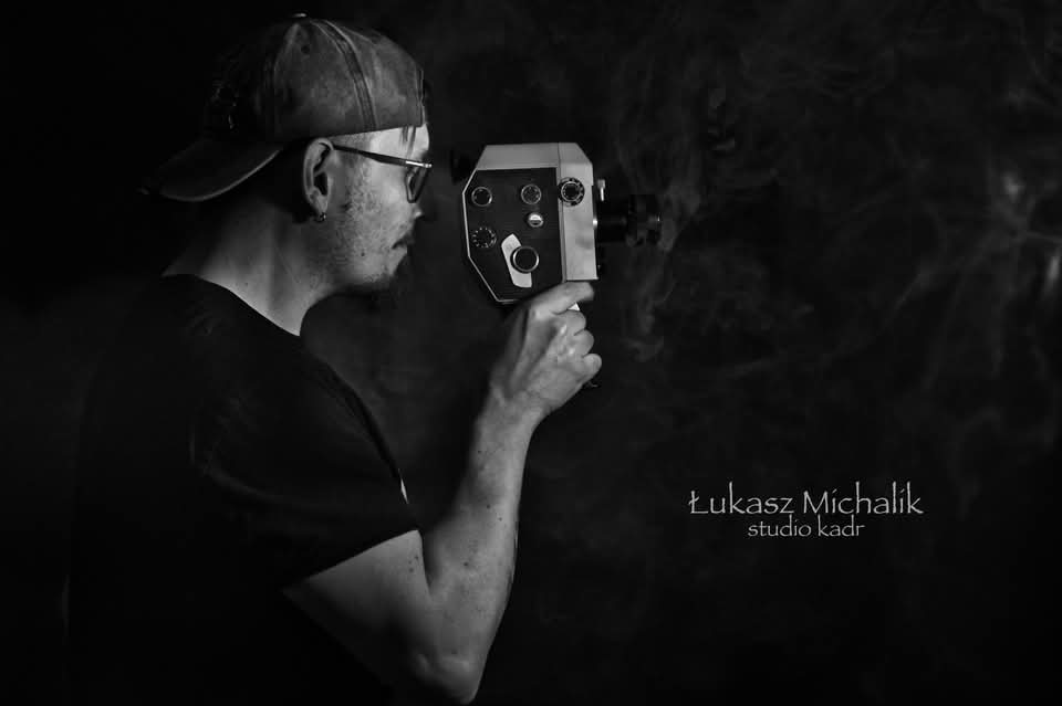 Czarno-białe zdjęcie mężczyzny z brodą i okularami, trzymającego starą kamerę filmową w Ustroniu. Stylizowane ujęcie z dymem w tle i logo studia.