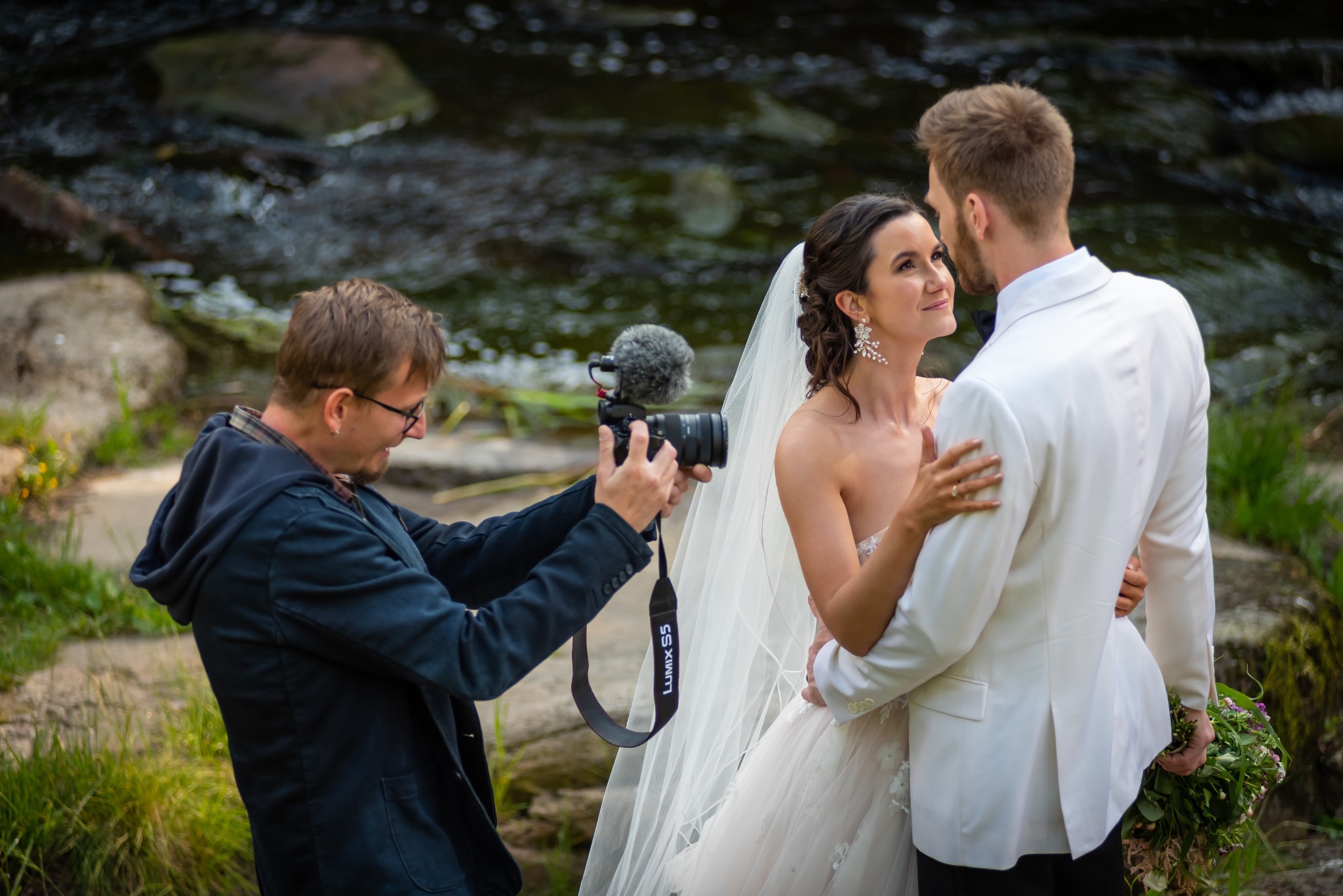Kamerzysta filmuje parę młodą na tle strumienia. Ujęcie z boku, widoczny mikrofon na kamerze Lumix S5. Panna młoda w sukni ślubnej, pan młody w białym smokingu.