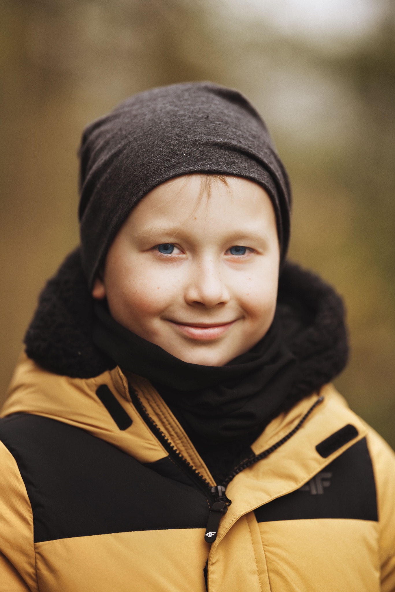 Portret uśmiechniętego chłopca o niebieskich oczach w czapce i kurtce 4F na rozmytym tle. Ujęcie z bliska, naturalne światło.