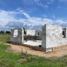 Marek Kruszczyński - Budowa domu z białych bloczków: wzniesione ściany parteru, widoczny pracownik wewnątrz, drewniane szalunki fundamentów, błękitne niebo z chmurami w tle.