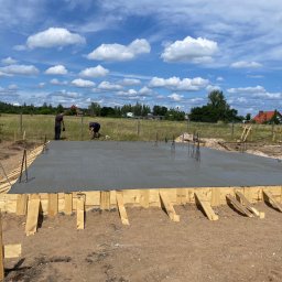 Marek Kruszczyński - Zalany fundament pod budowę domu, drewniane szalunki, wystające pręty zbrojeniowe, dwóch pracowników w tle, błękitne niebo z chmurami.